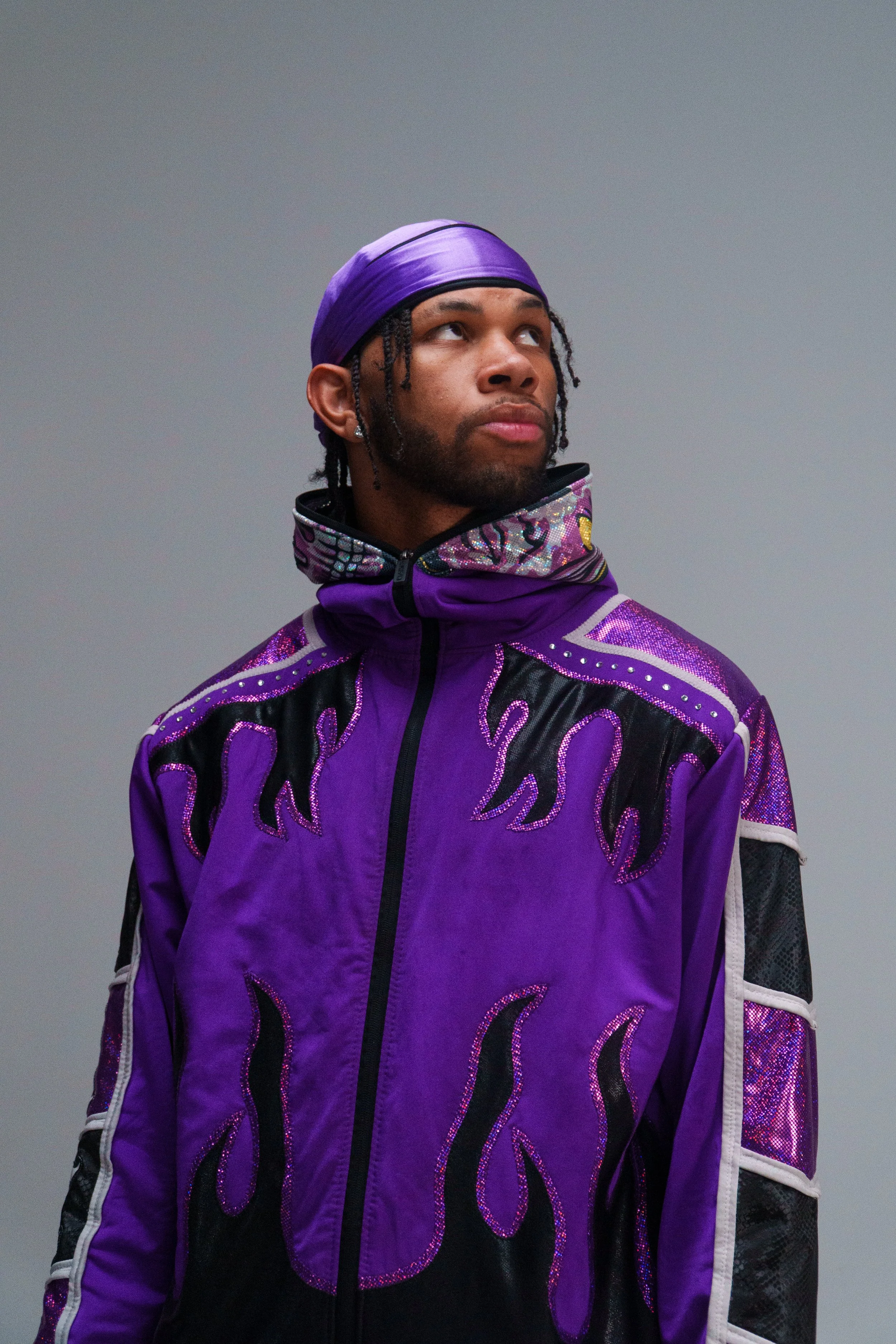 Man wearing a purple jacket with flame designs and a matching purple headscarf, looking to the side against a gray background.