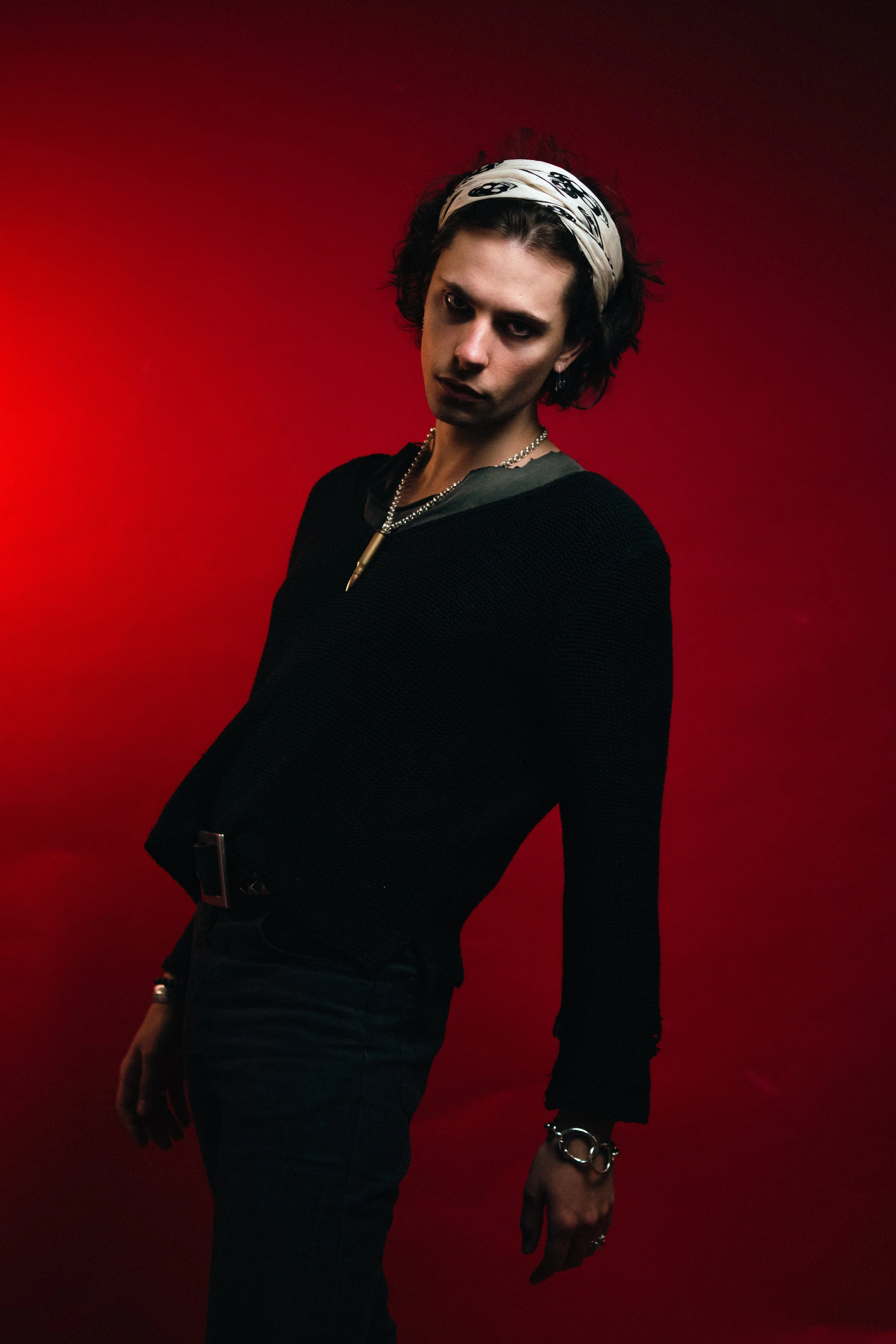 A person with short, curly black hair wearing a white bandana, dark eye makeup, black clothing, and jewelry, posing against a red background.