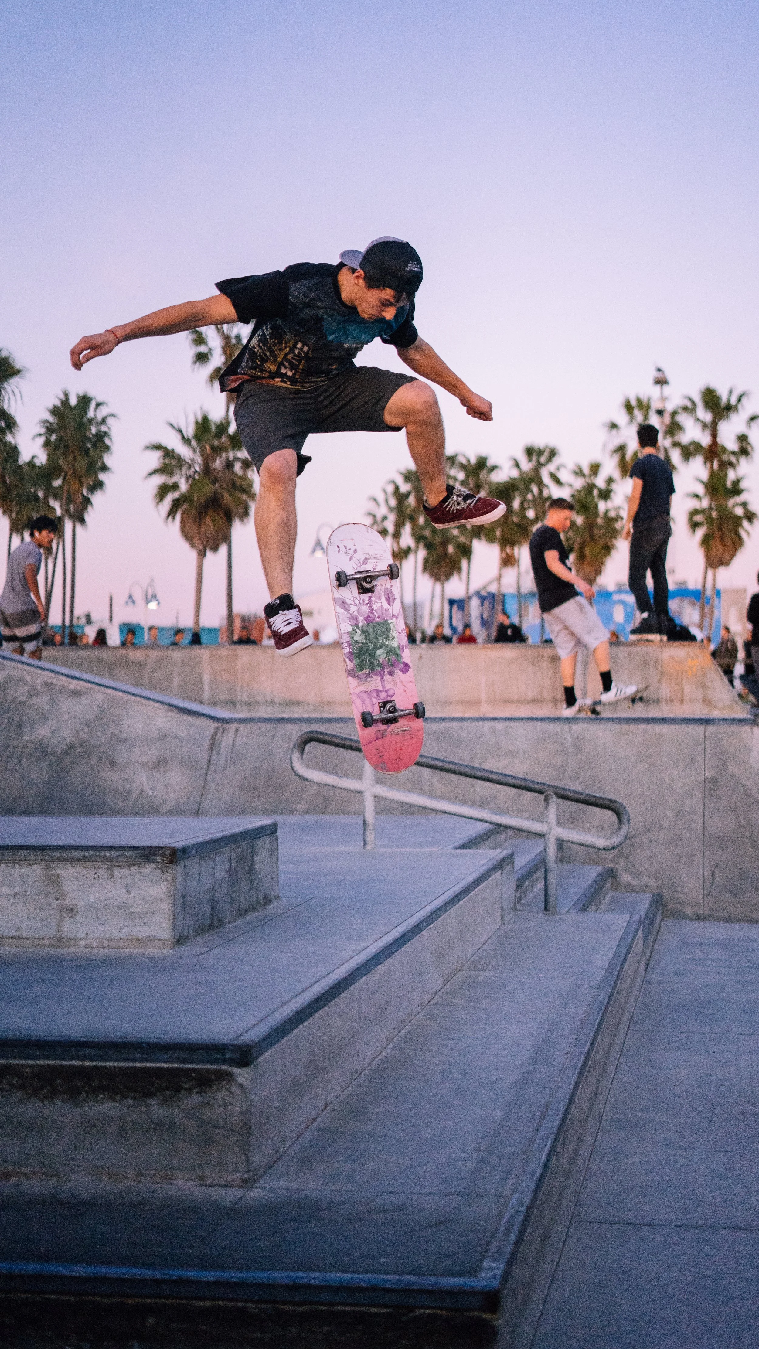Skateboarder performing a trick on a rail at a skate park during sunset, with palm trees and other skaters in the background.
