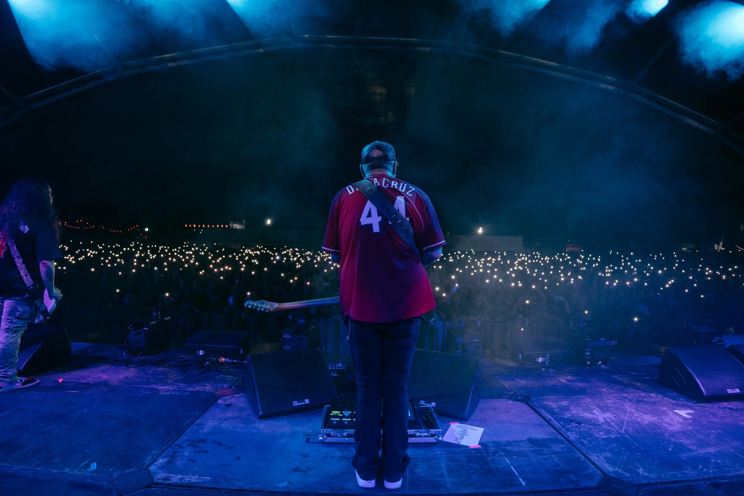 A musician on stage during a concert, facing a large audience with many lights visible in the dark. The musician is wearing a red jersey and a cap, with a guitar strap across the back.