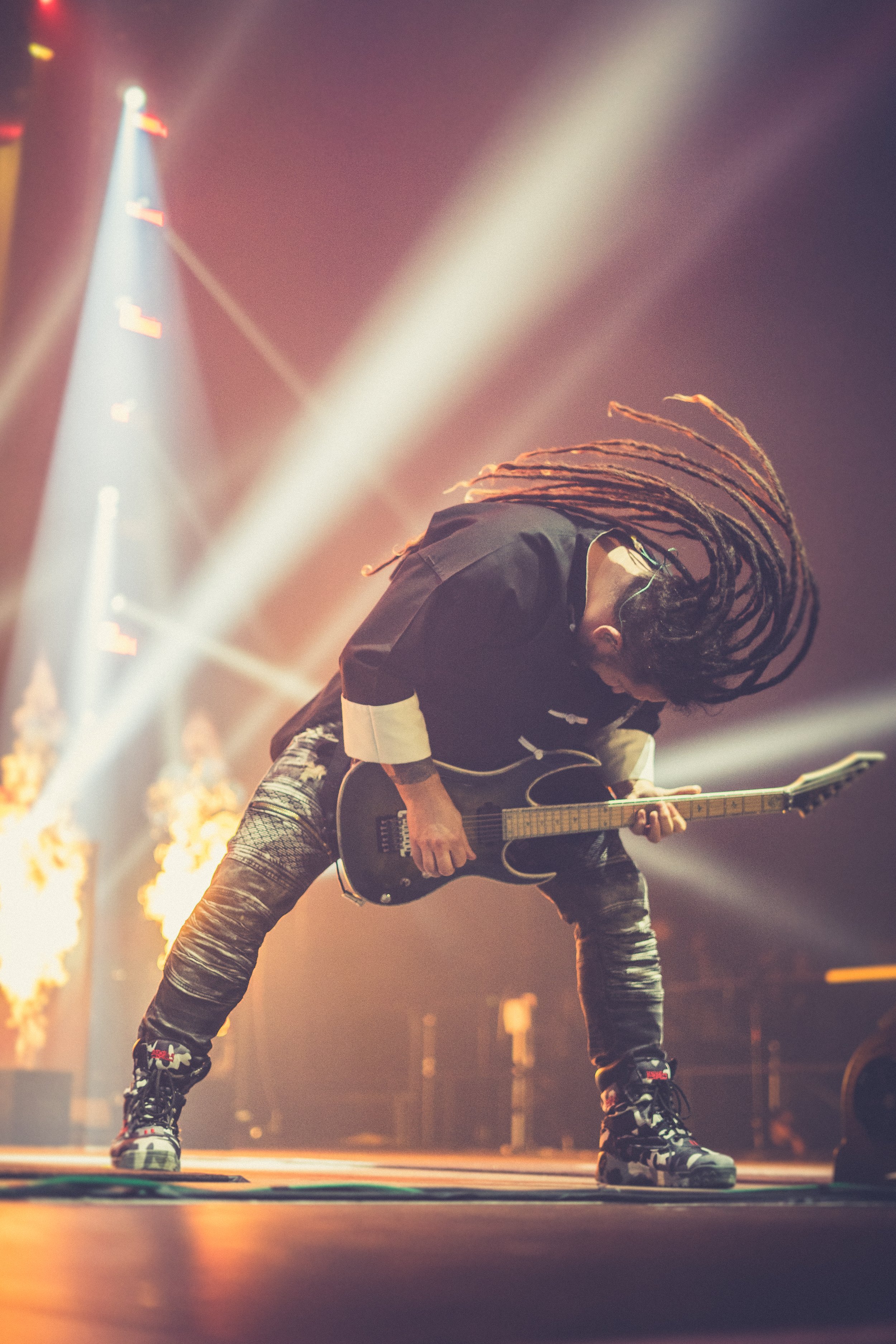 A musician with long dreadlocks playing an electric guitar on stage with bright lights and flames in the background.