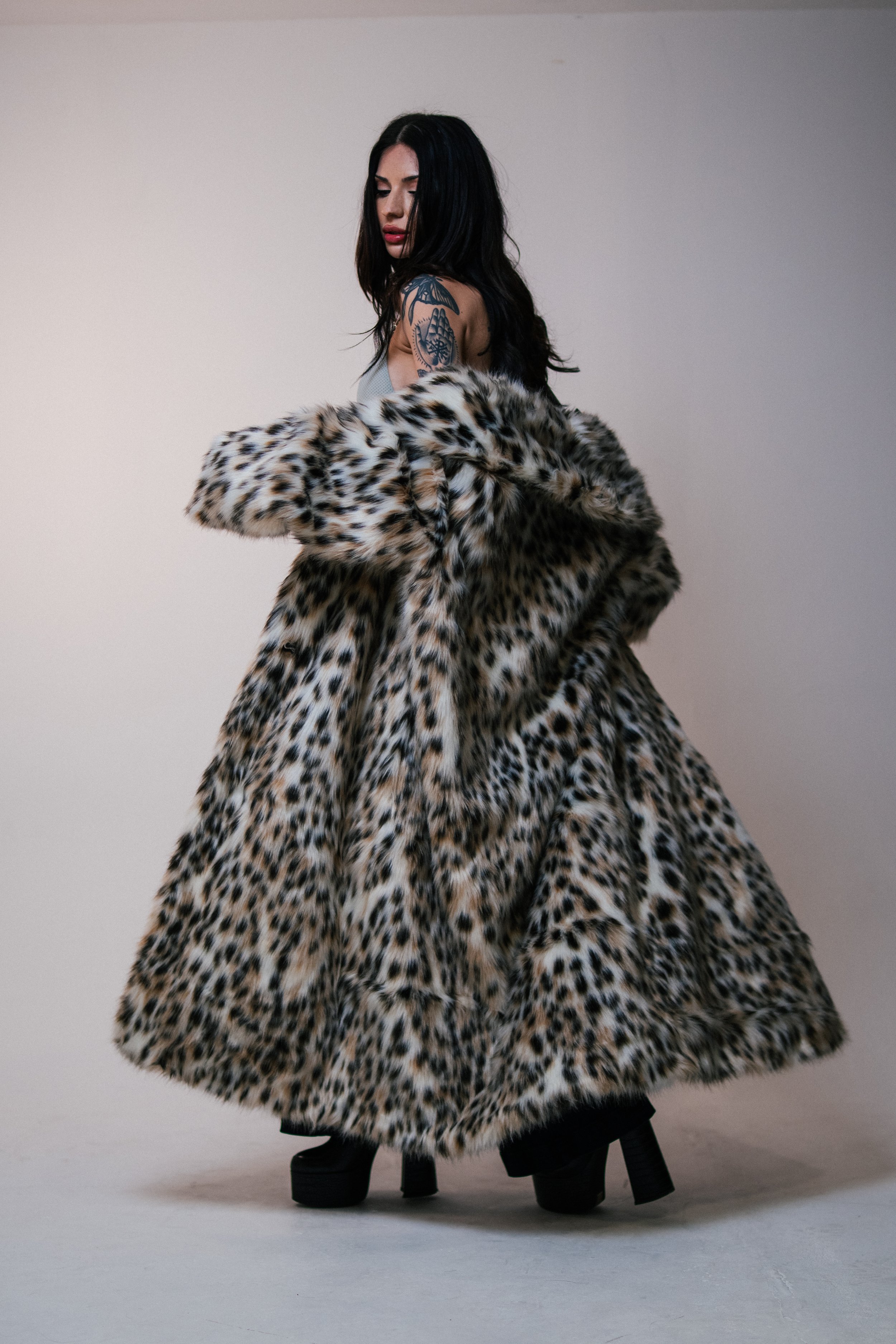 A woman with black hair and tattoos wearing a leopard-print fur coat and black platform shoes, standing against a plain light background.
