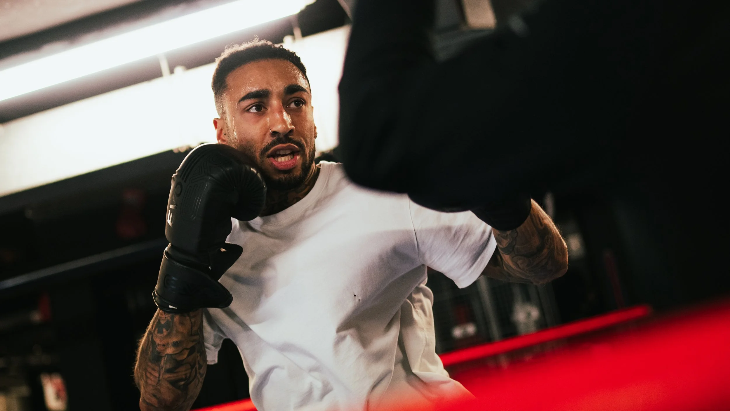 A male boxer with tattoos on his arms wearing black boxing gloves and a white T-shirt practicing a punch in a gym.