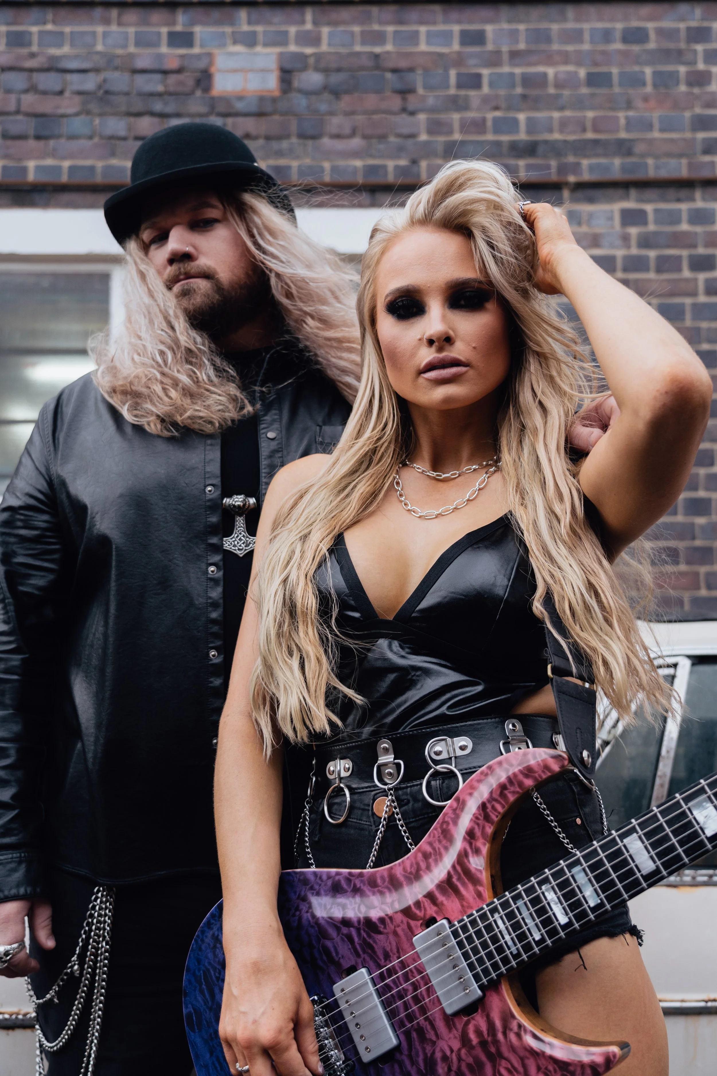Two musicians posing outdoors; a woman in the front holds a purple and pink electric guitar, and a man stands behind her. They style with edgy, rebellious fashion, with the woman wearing a black leather top, chains, and long wavy blonde hair, and the