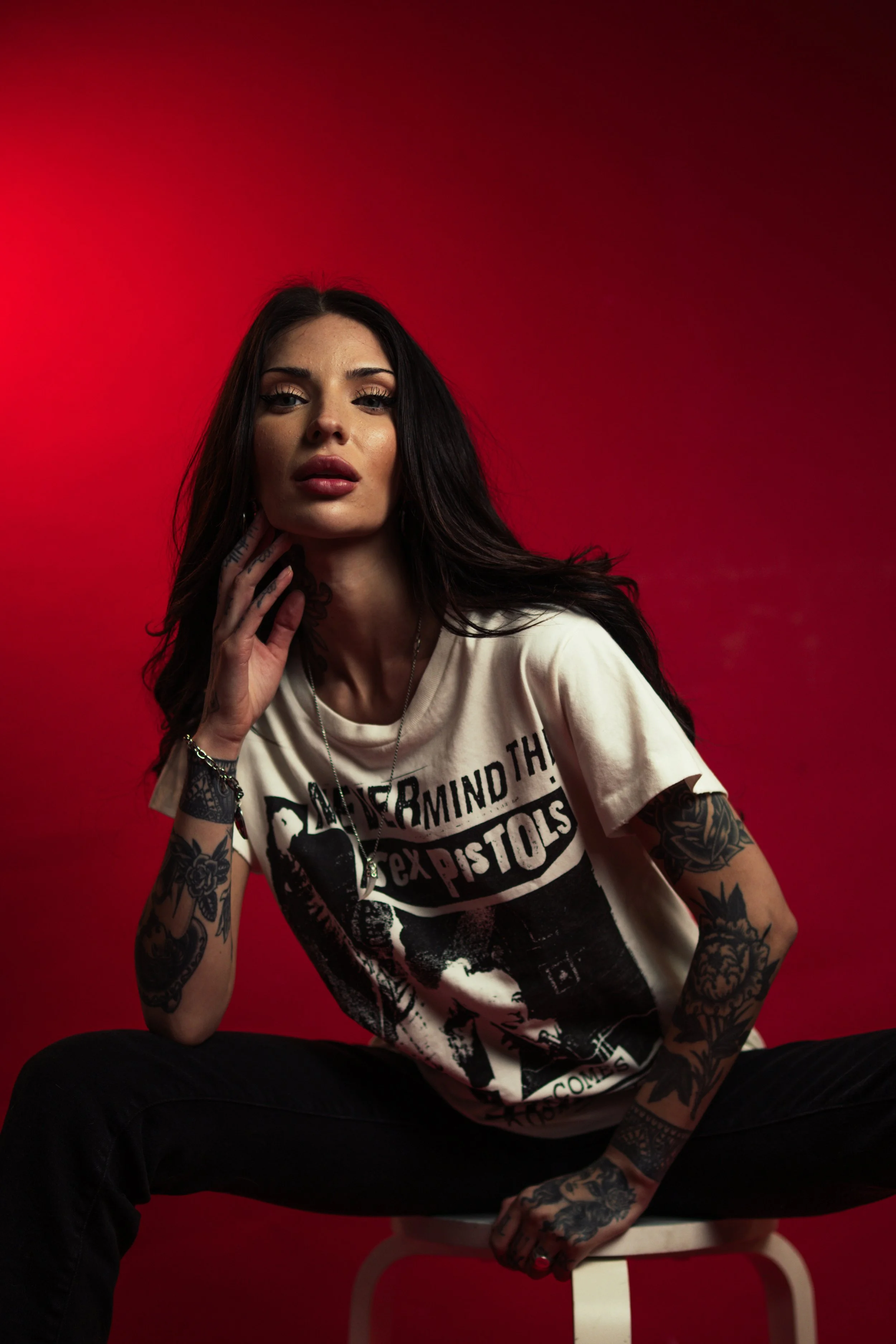A woman with long dark hair and tattoos on her arms, wearing a graphic T-shirt, sits on a stool against a red background.