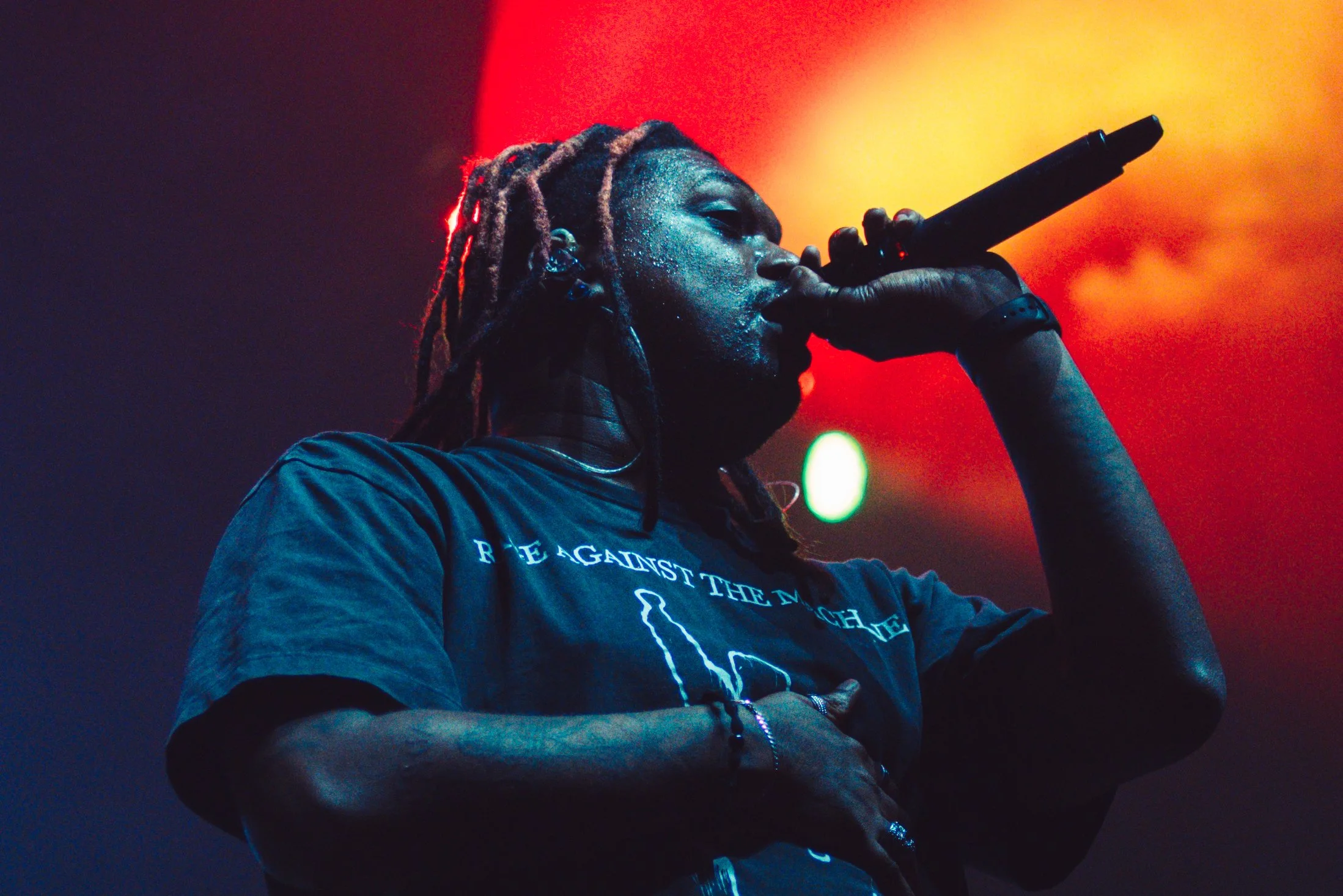 A male rapper with dreadlocks performing on stage, holding a microphone close to his mouth, with colorful stage lighting in the background.