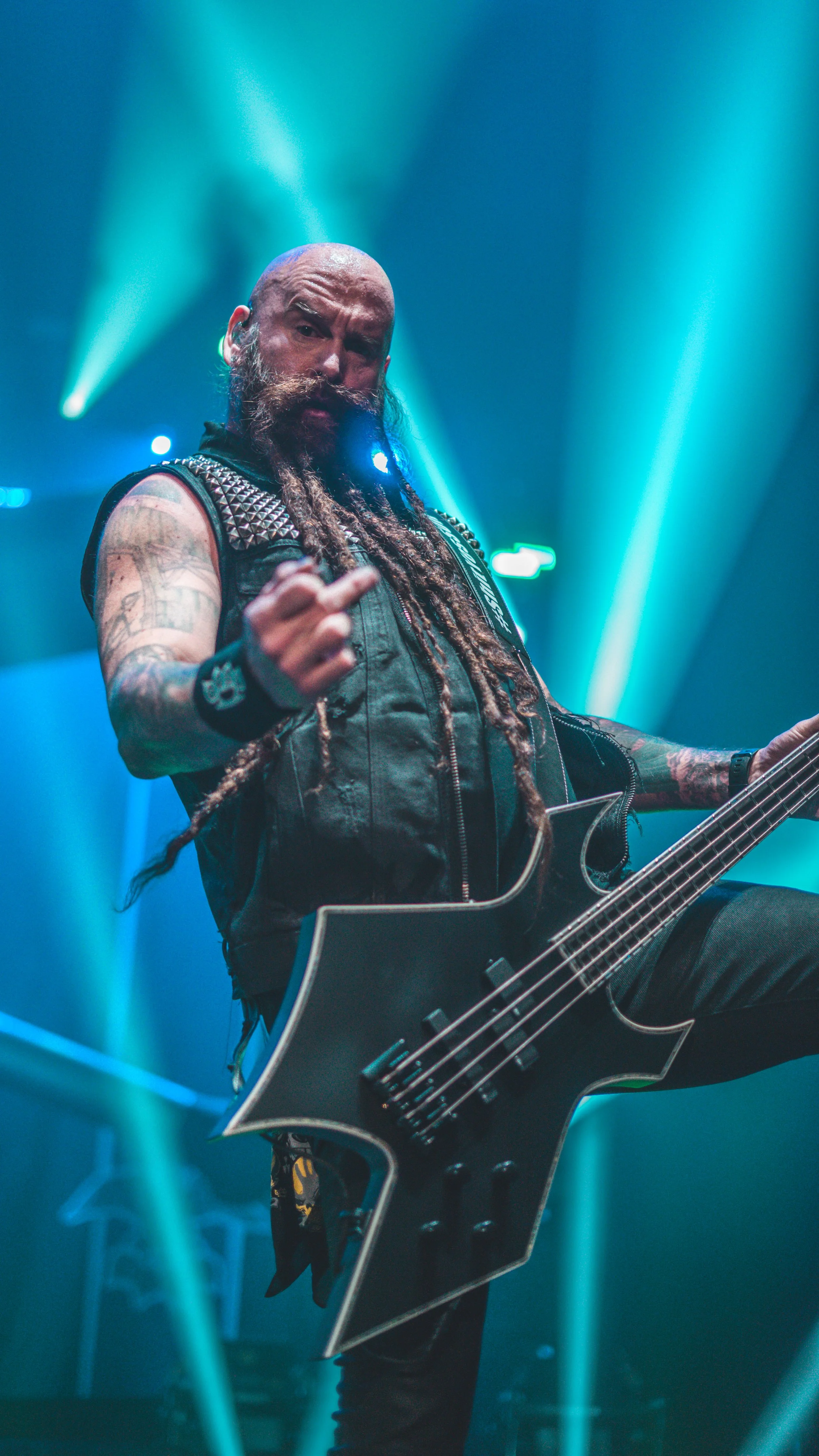 A male musician with a beard and tattoos playing a black electric bass guitar on stage, making a hand gesture, with bright blue stage lights in the background.