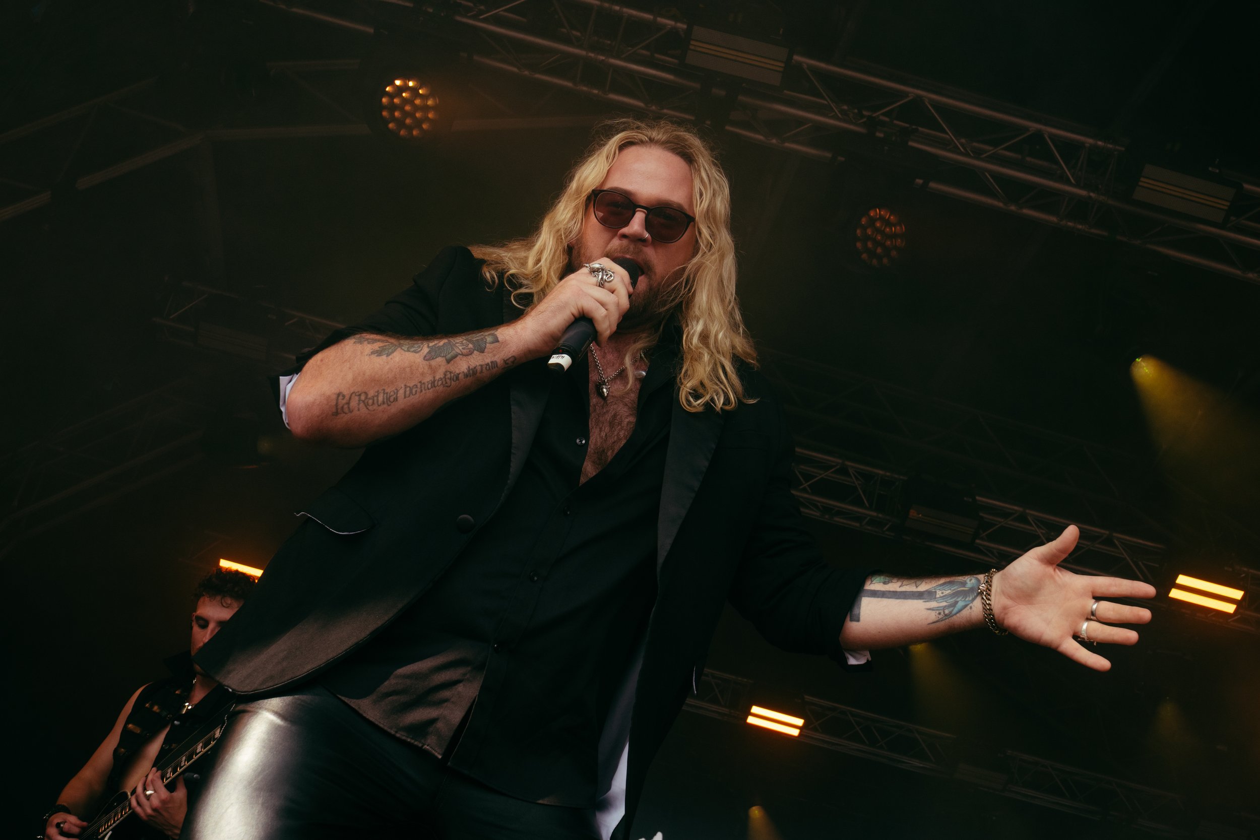 A male singer with long blonde hair, sunglasses, and tattoos performs on stage with a microphone in hand, wearing a black suit jacket and shirt, during a concert plus a guitarist in the background.