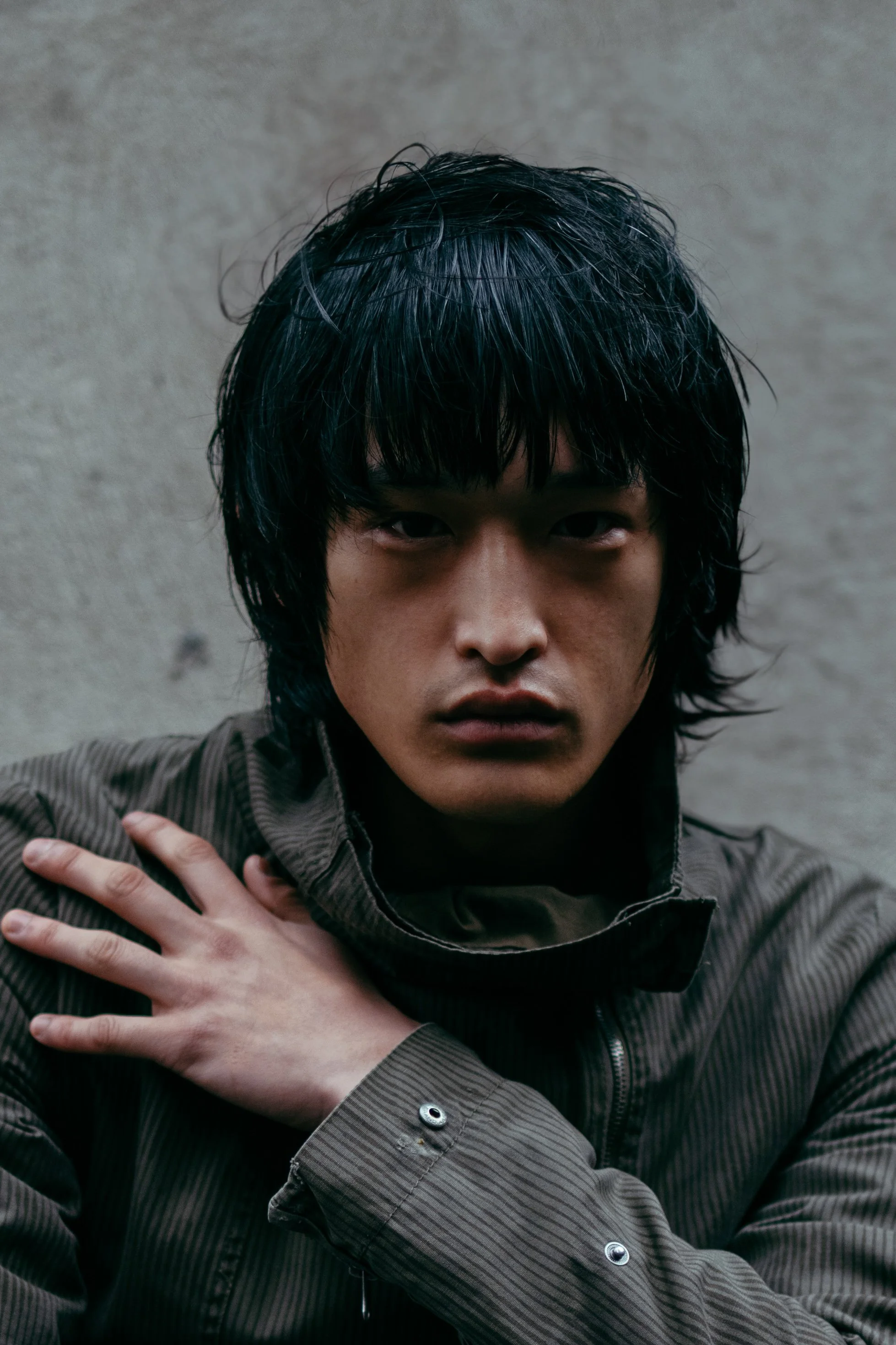 A young man with black hair looks directly at the camera, wearing a brown jacket, with one hand on his shoulder, against a plain, textured background.