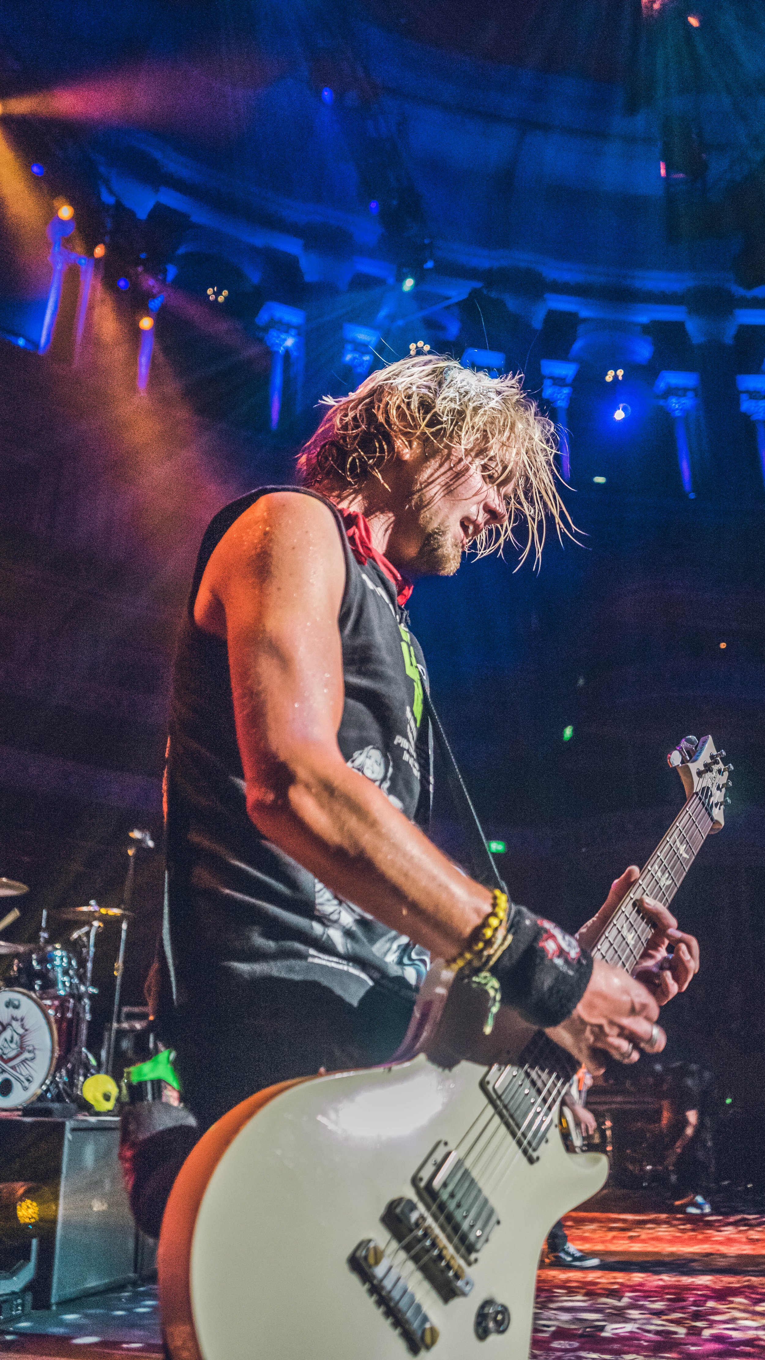 A male guitarist performing on stage with colorful lighting, wearing a sleeveless shirt and wristbands.