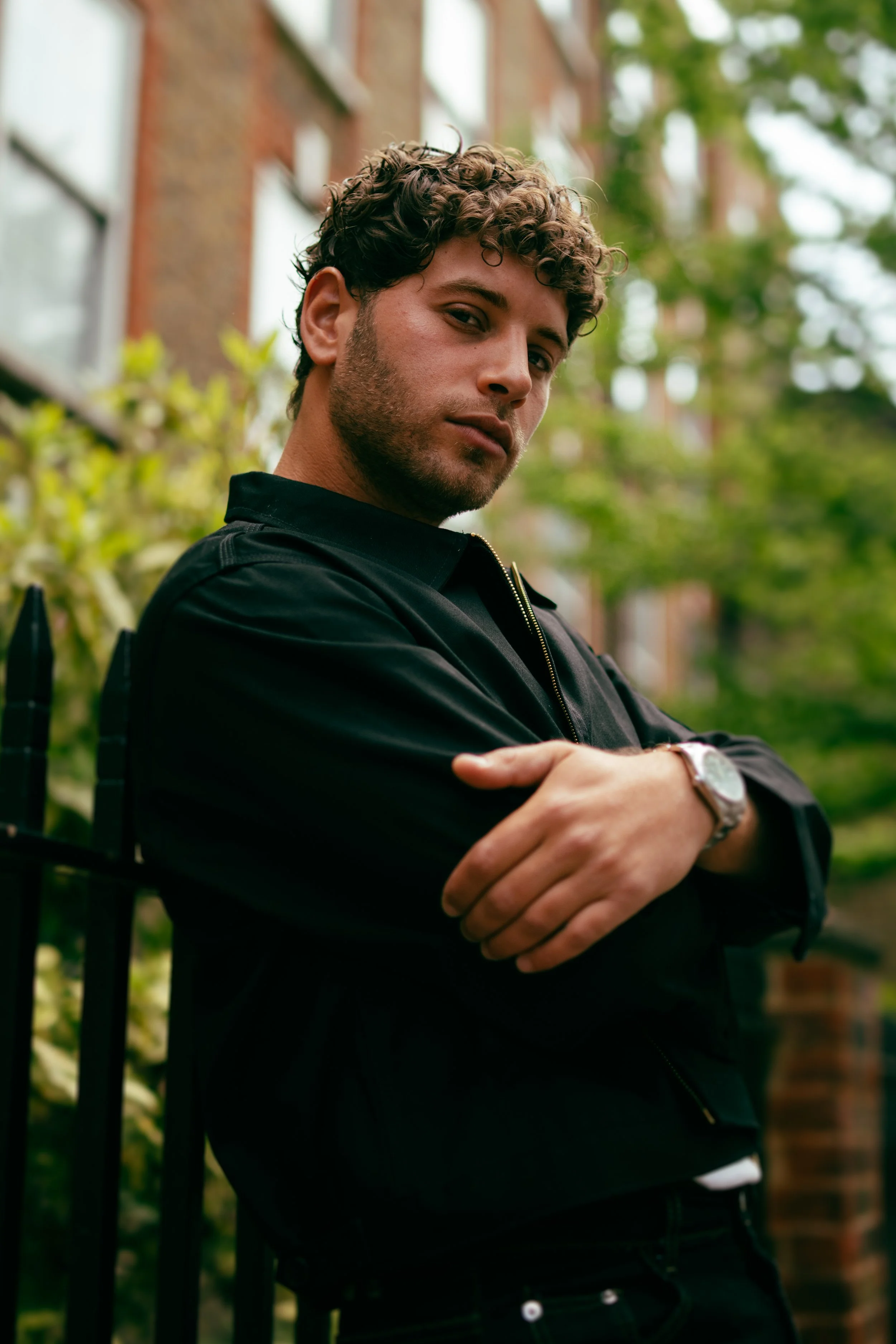 A young man with curly hair and a beard, wearing a black jacket and a watch, standing outside with greenery and brick buildings in the background, crossing his arms and looking at the camera.