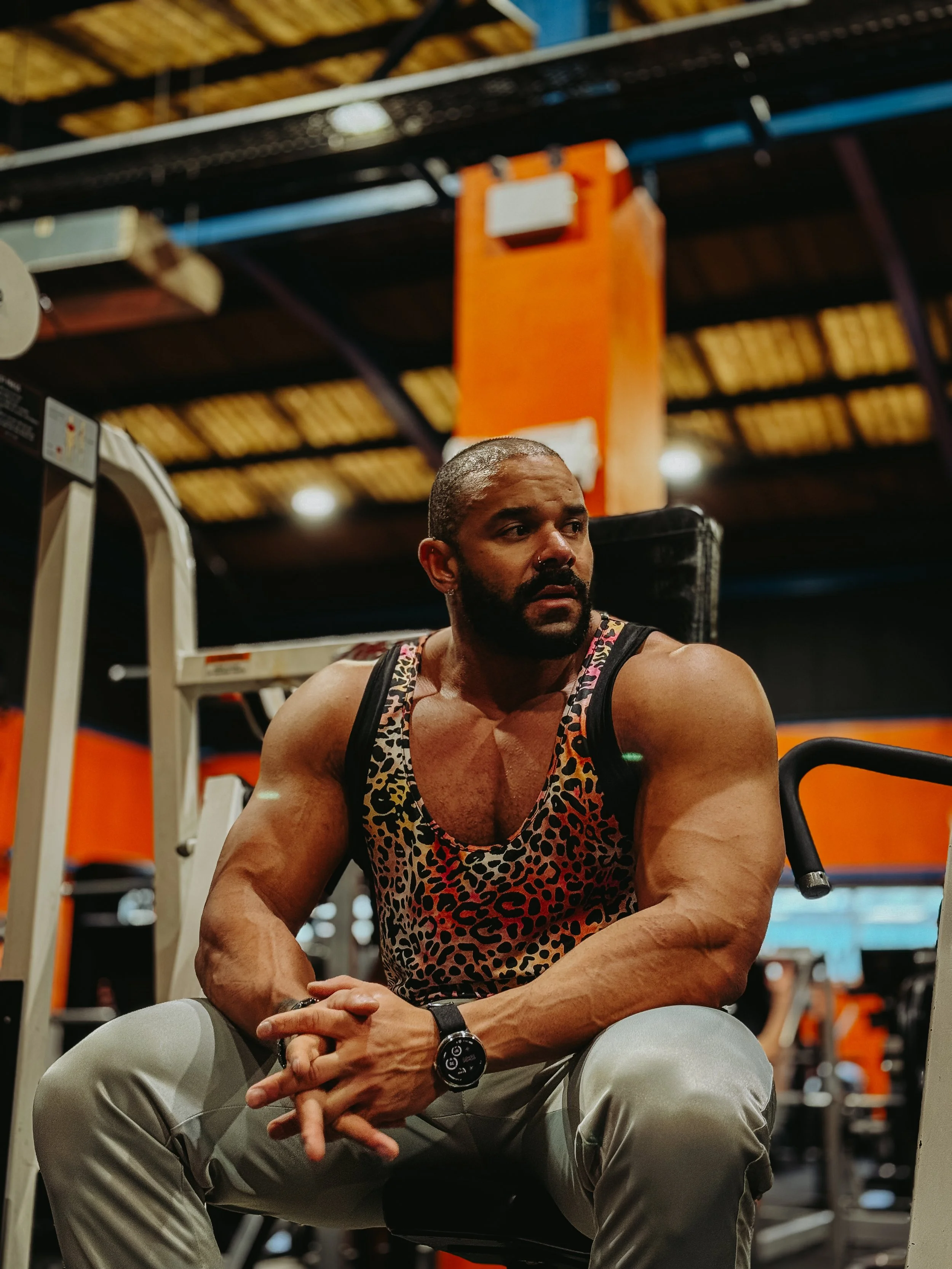 A muscular man sitting in a gym, wearing a leopard print tank top and light-colored pants, with a serious expression, surrounded by gym equipment.