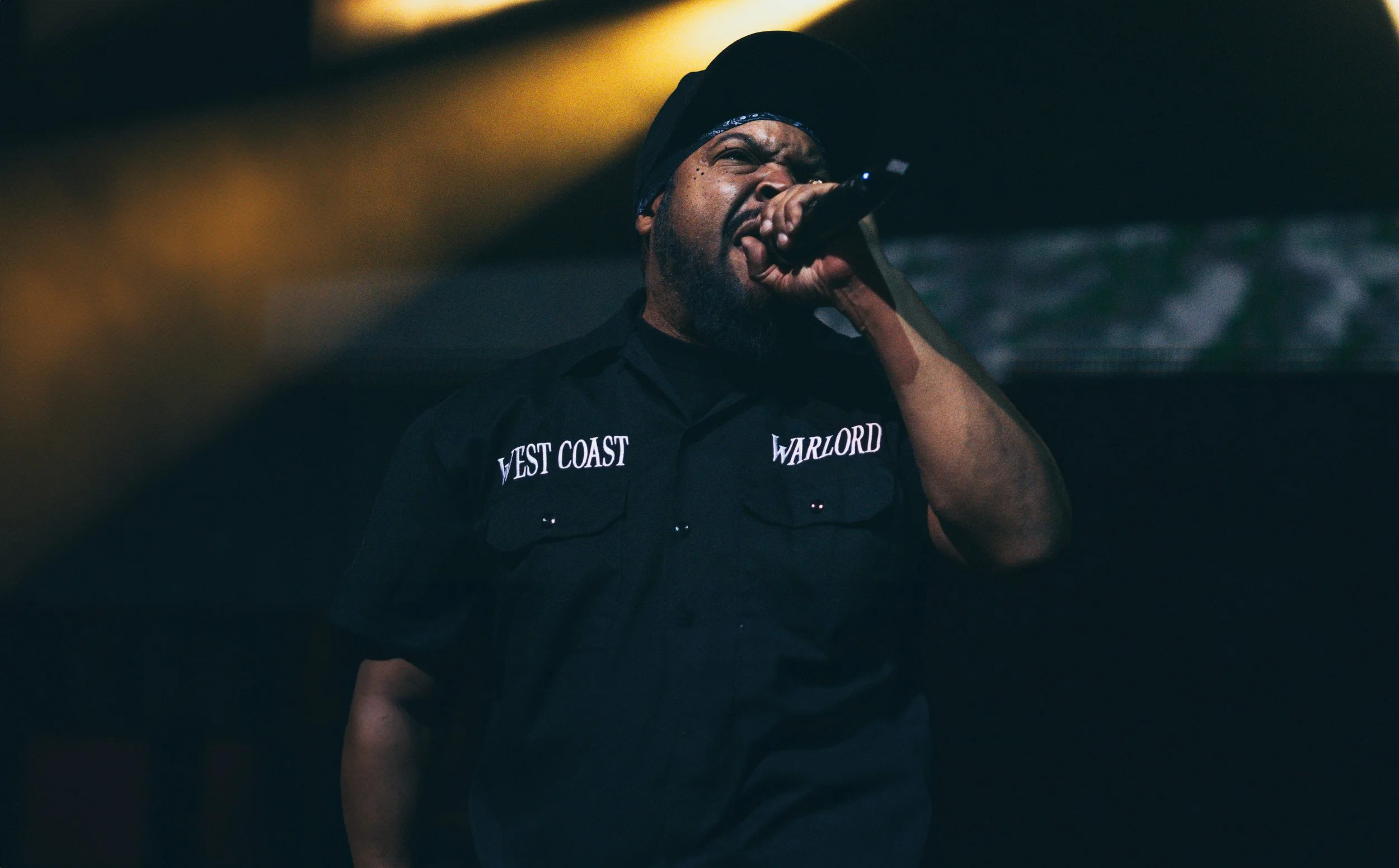 A man wearing a black shirt with the words 'VEST COAST' and 'WARLORD' singing into a microphone on stage.