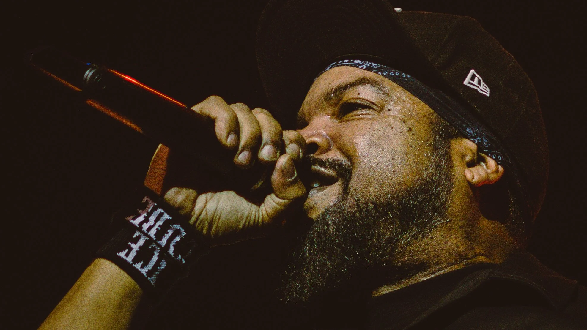A man with a beard singing into a microphone, wearing a black hat and a wristband, with a dark background.