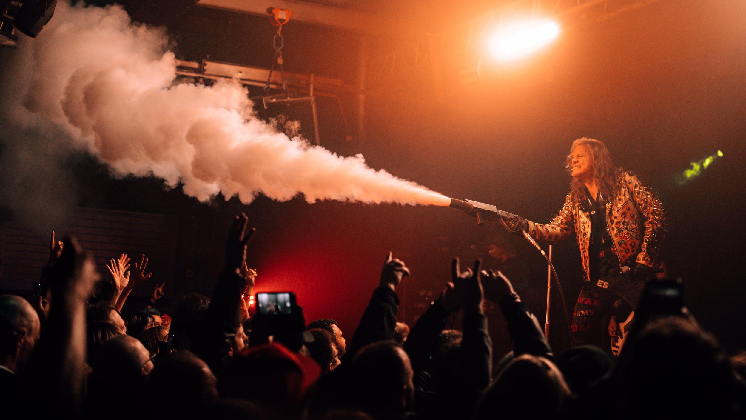A performer holding a smoke emitter on stage, emitting a thick cloud of white smoke toward the audience, who are raising their hands and taking pictures in a concert setting with warm lighting.