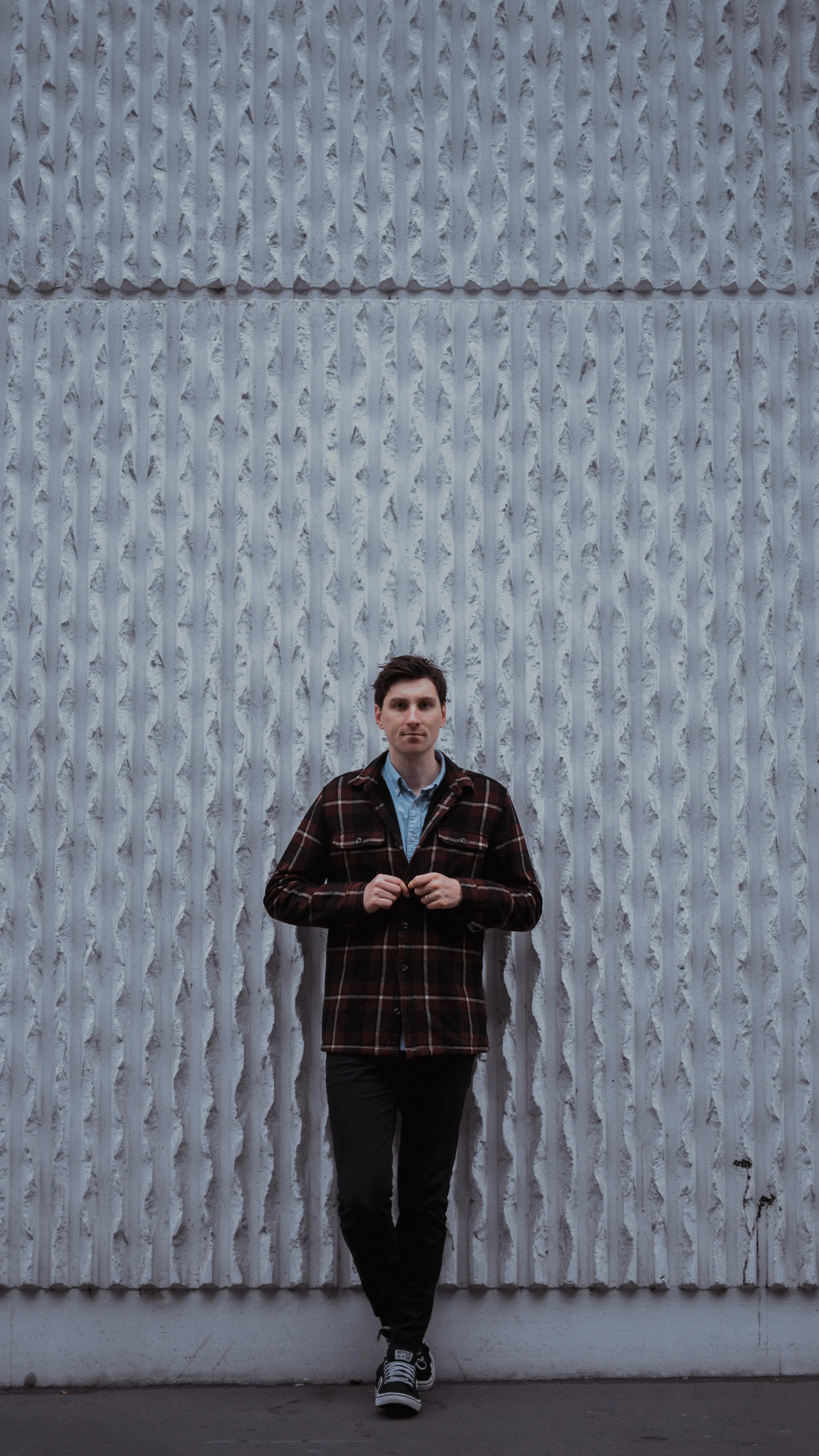 A young man with dark hair standing in front of a large textured concrete wall, wearing a plaid jacket over a blue shirt and black pants.