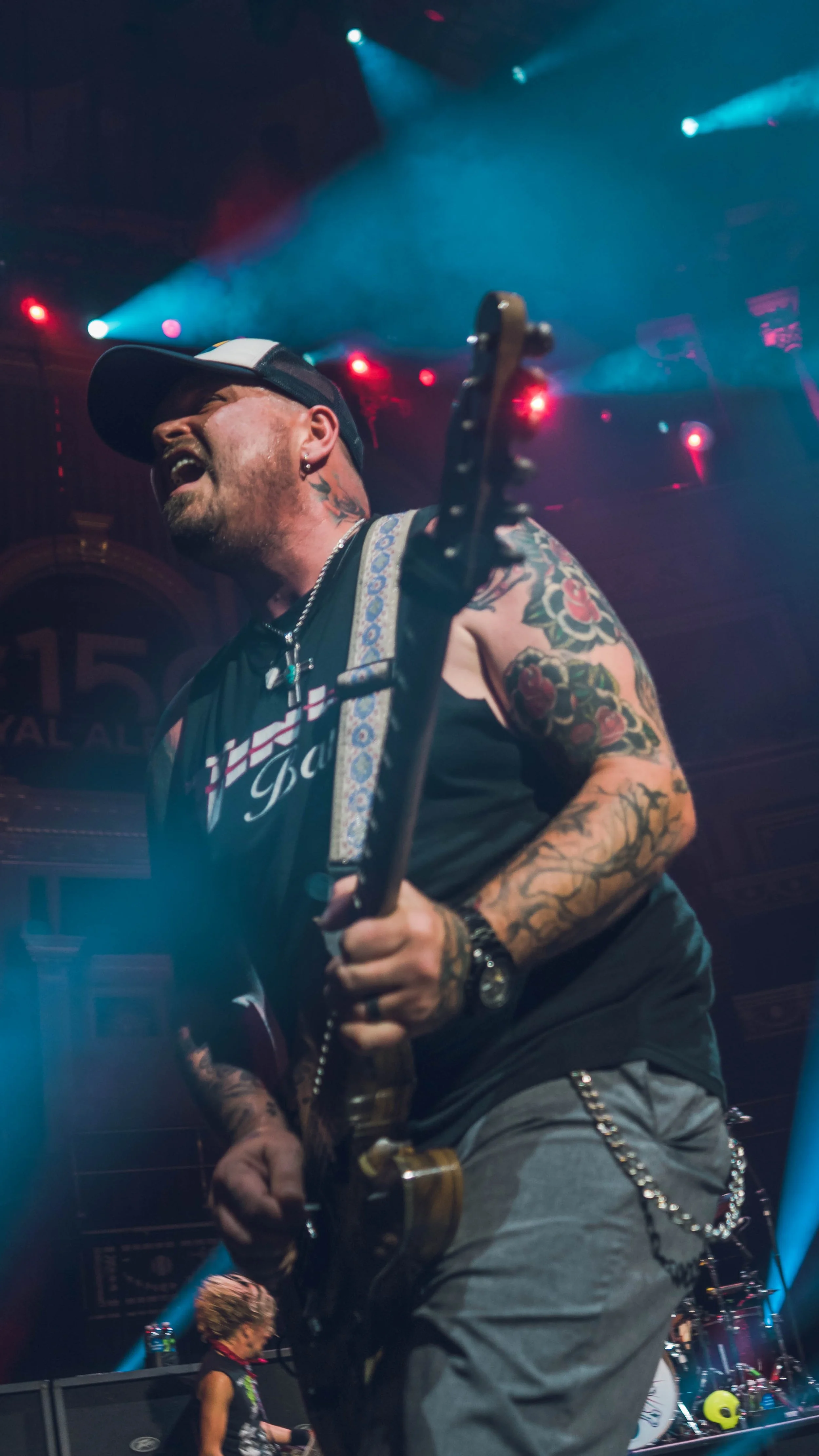 Male guitarist performing on stage with tattoos, wearing a hat and sleeveless shirt, with colorful stage lights overhead.