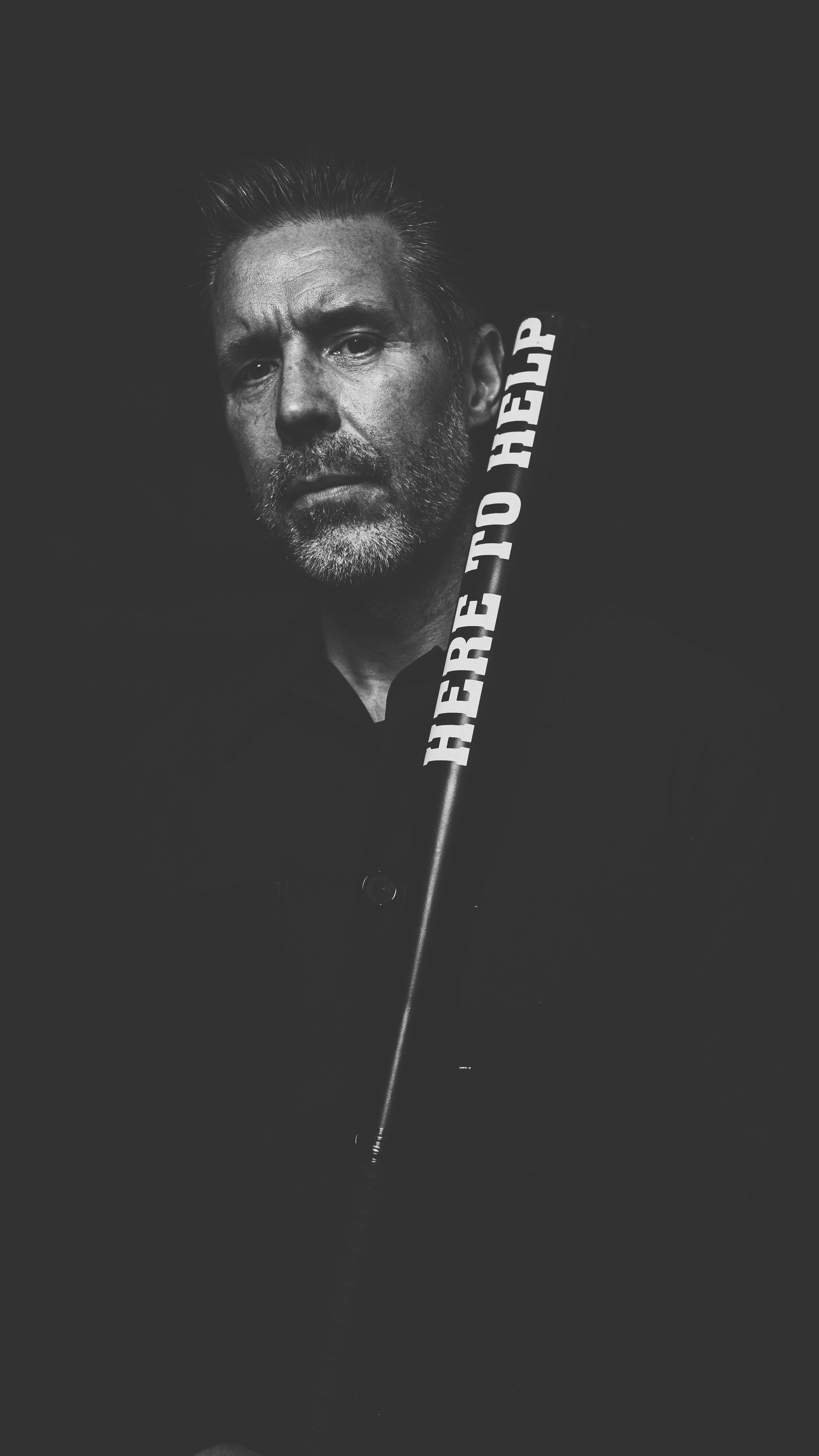 Black and white portrait of a middle-aged man with a beard, holding a baseball bat with the words 'HERE TO HELP' written on it, against a dark background.
