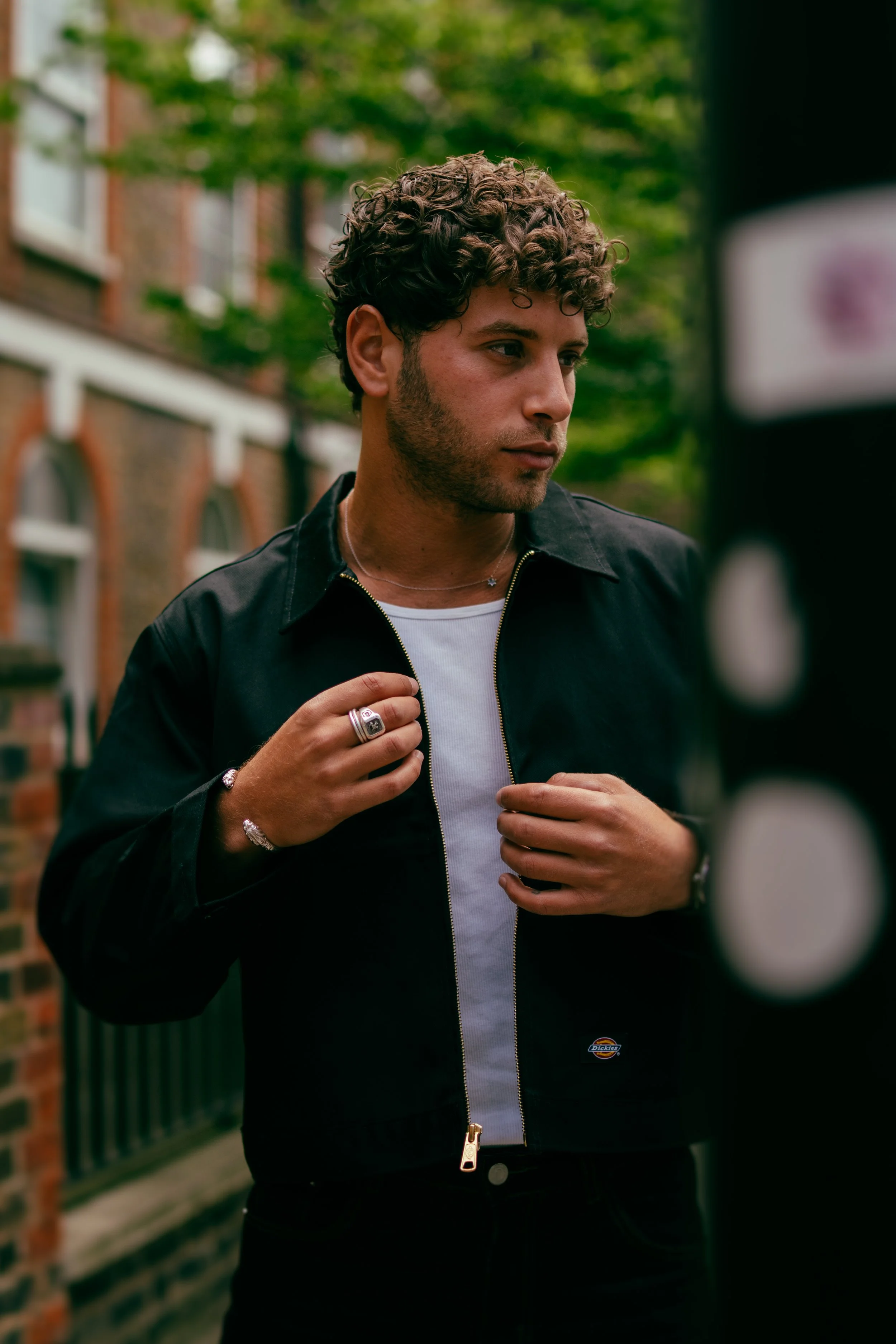 A young man with curly brown hair and a slight beard adjusts his black jacket while standing outdoors in front of a brick building with green trees in the background.