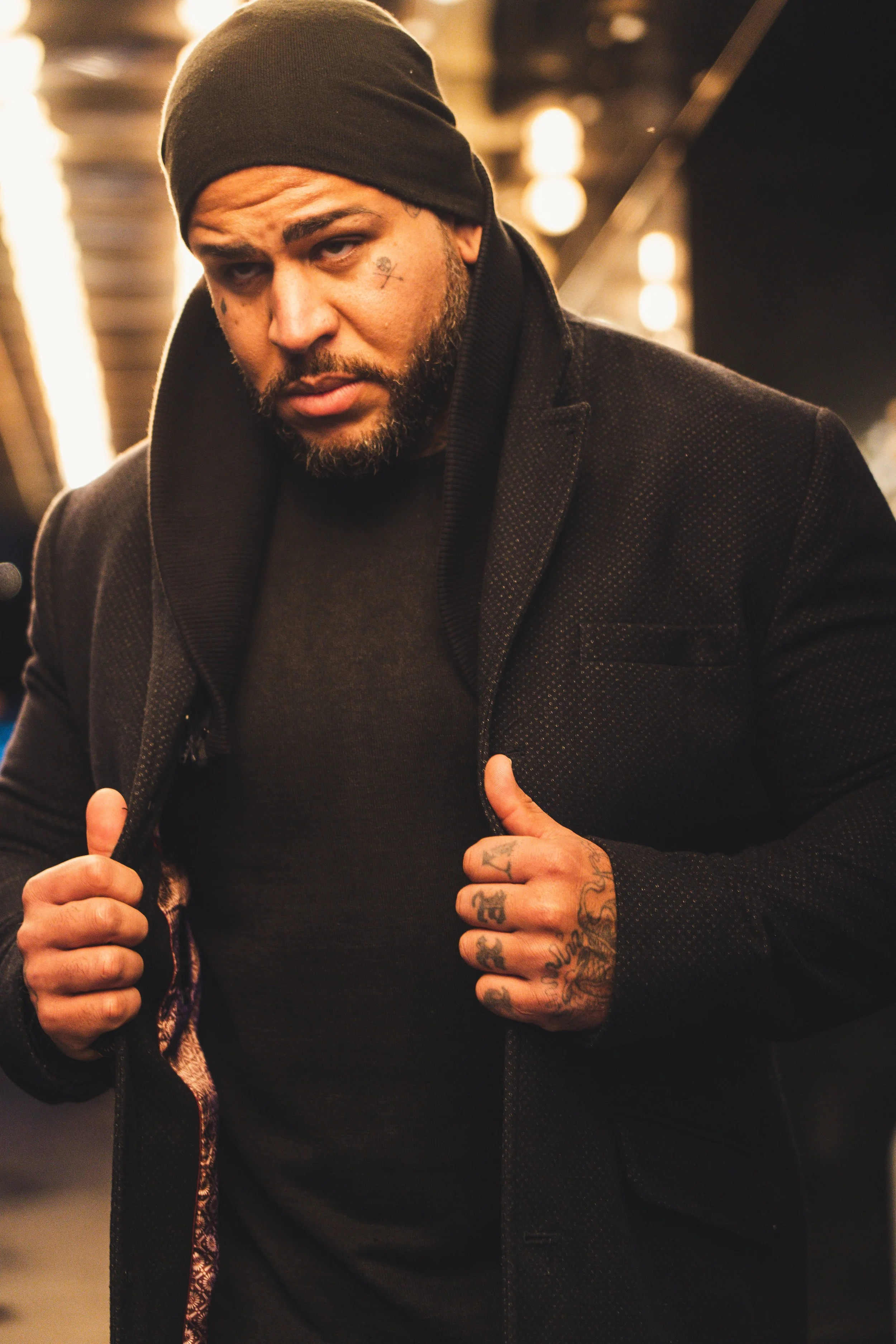 A man with tattoos on his hands and face is wearing a black beanie, black shirt, and black jacket, holding the lapels of his jacket with both hands and looking into the camera.