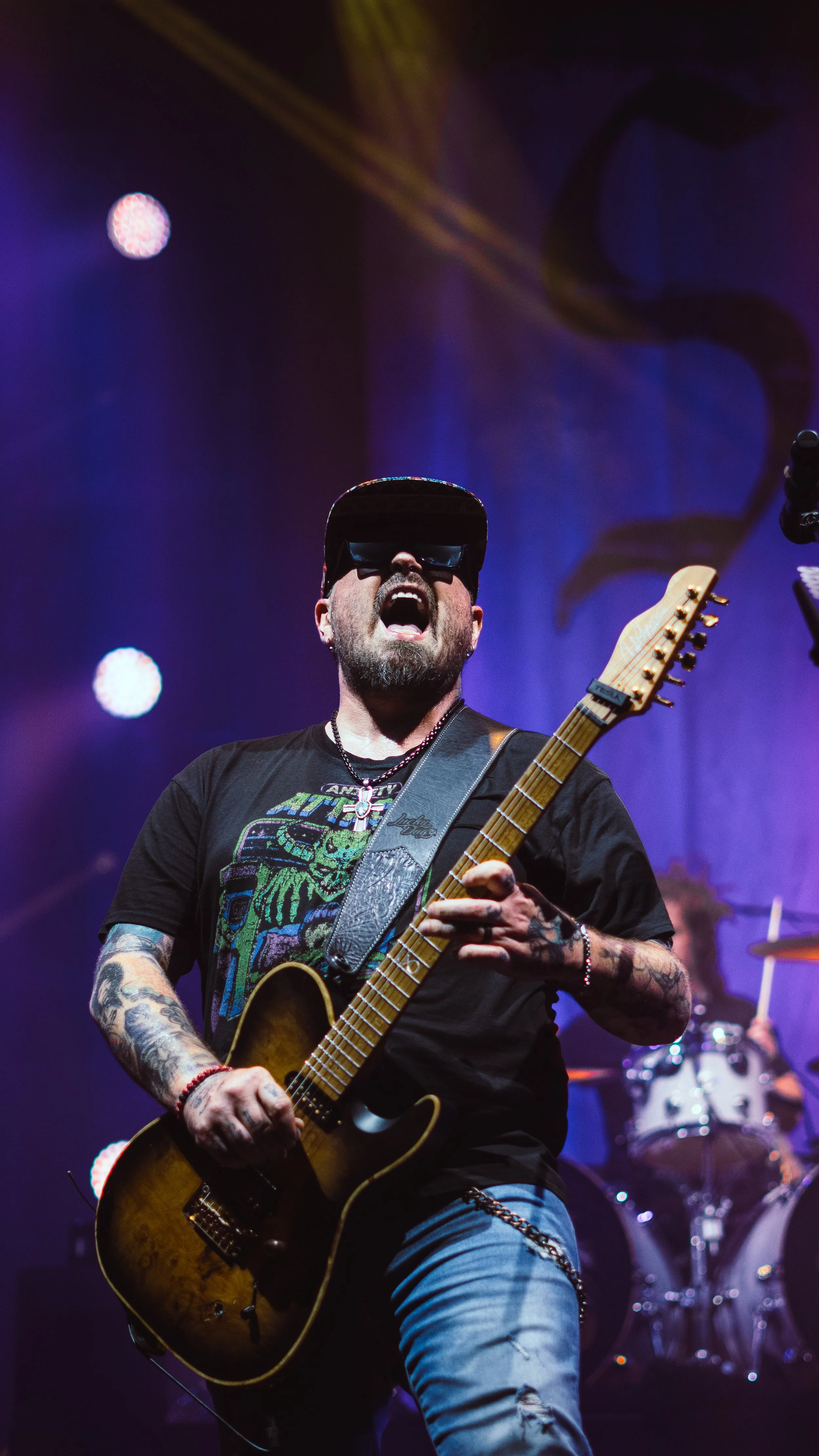 A man wearing sunglasses and a black cap, passionately playing an electric guitar on stage during a concert, illuminated by colorful stage lights.