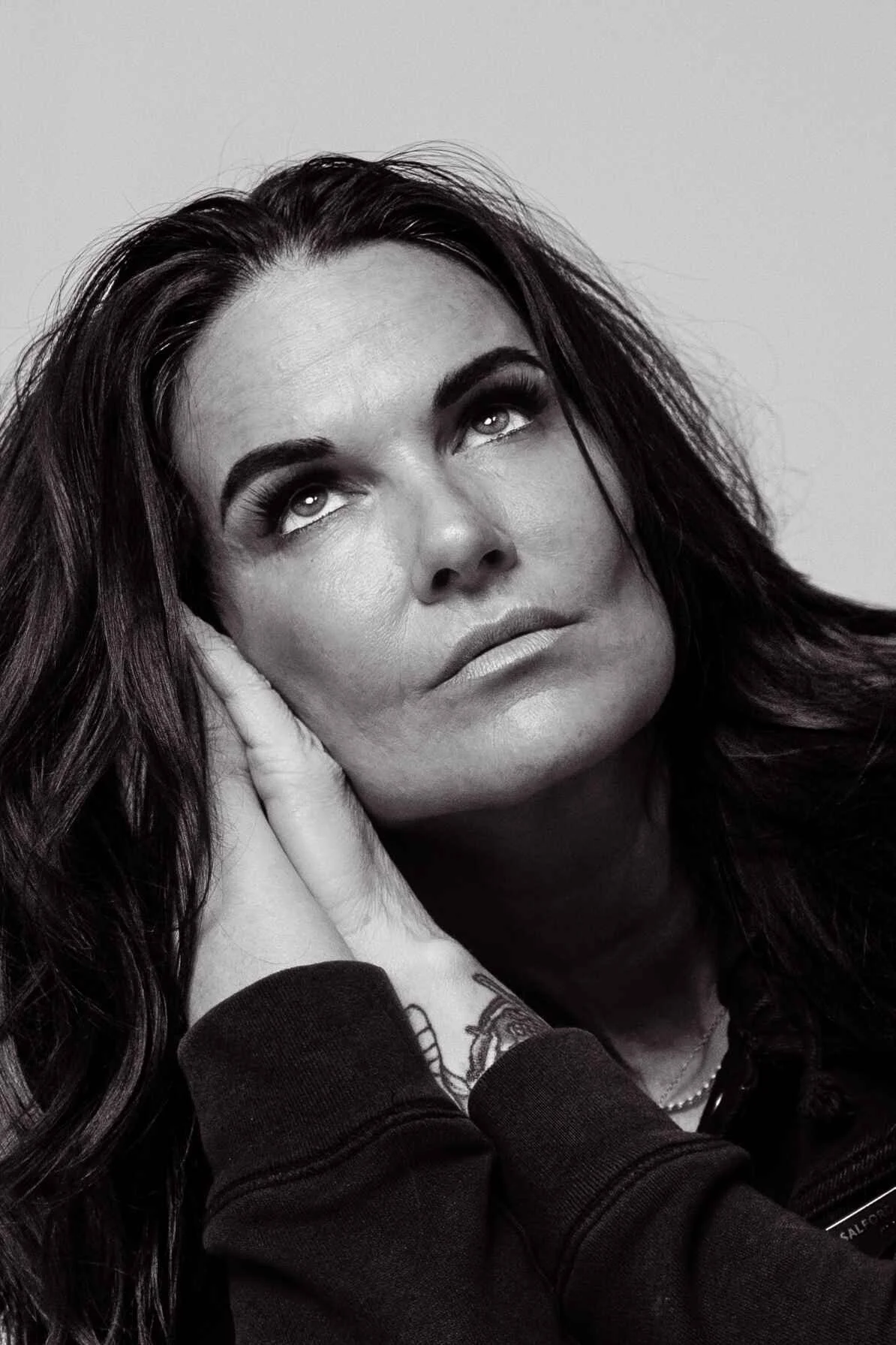 Black and white close-up of a woman with long dark hair, gazing upward with one hand resting on her face and the other on her neck, wearing a dark shirt.