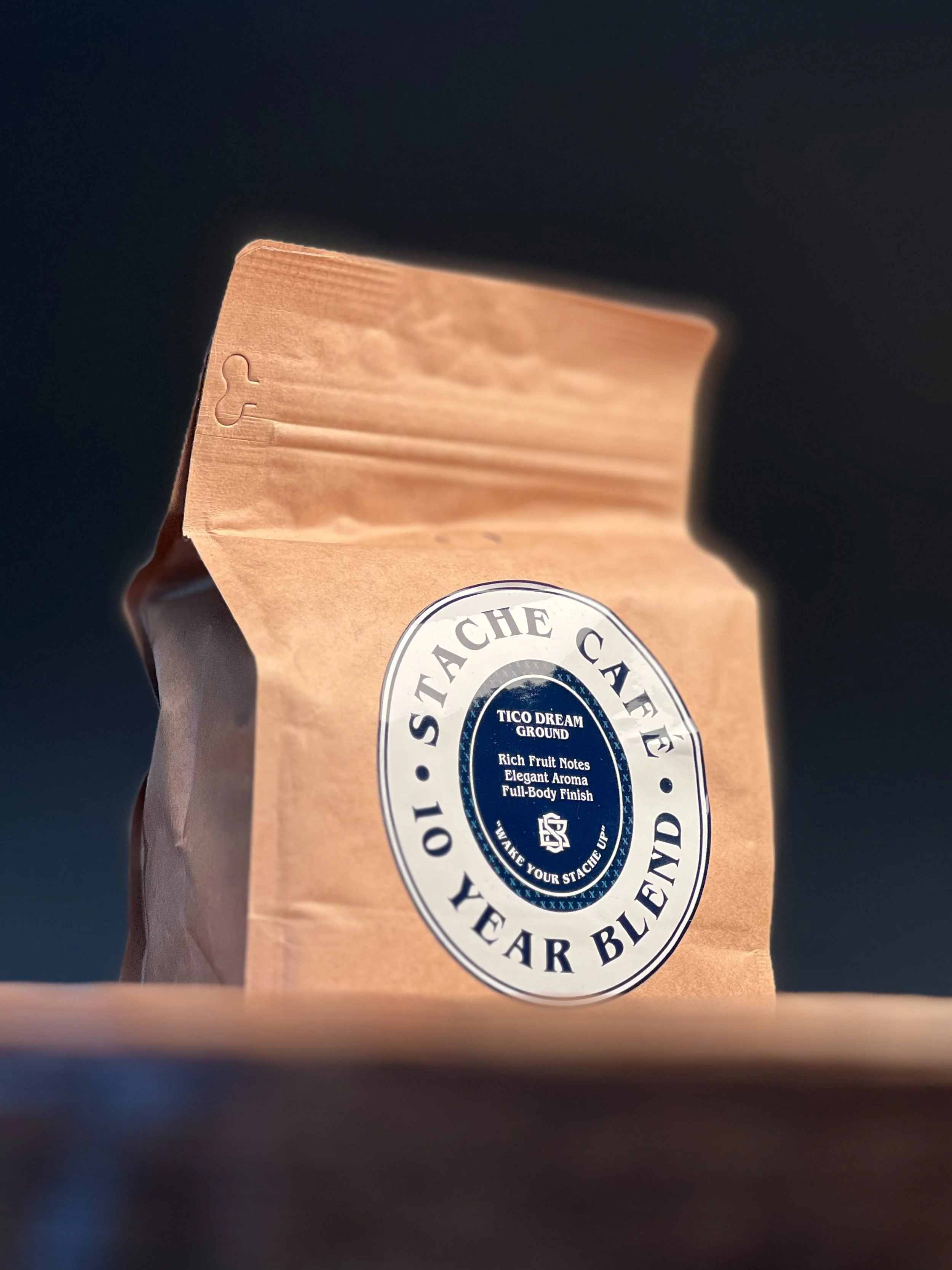 A brown paper bag of Stache Coffee with a round label that reads 'TICO DREAM GROUND' and describes rich fruit notes, elegant aroma, and full-body finish surrounding a logo and the phrase 'Wake your stache up'.