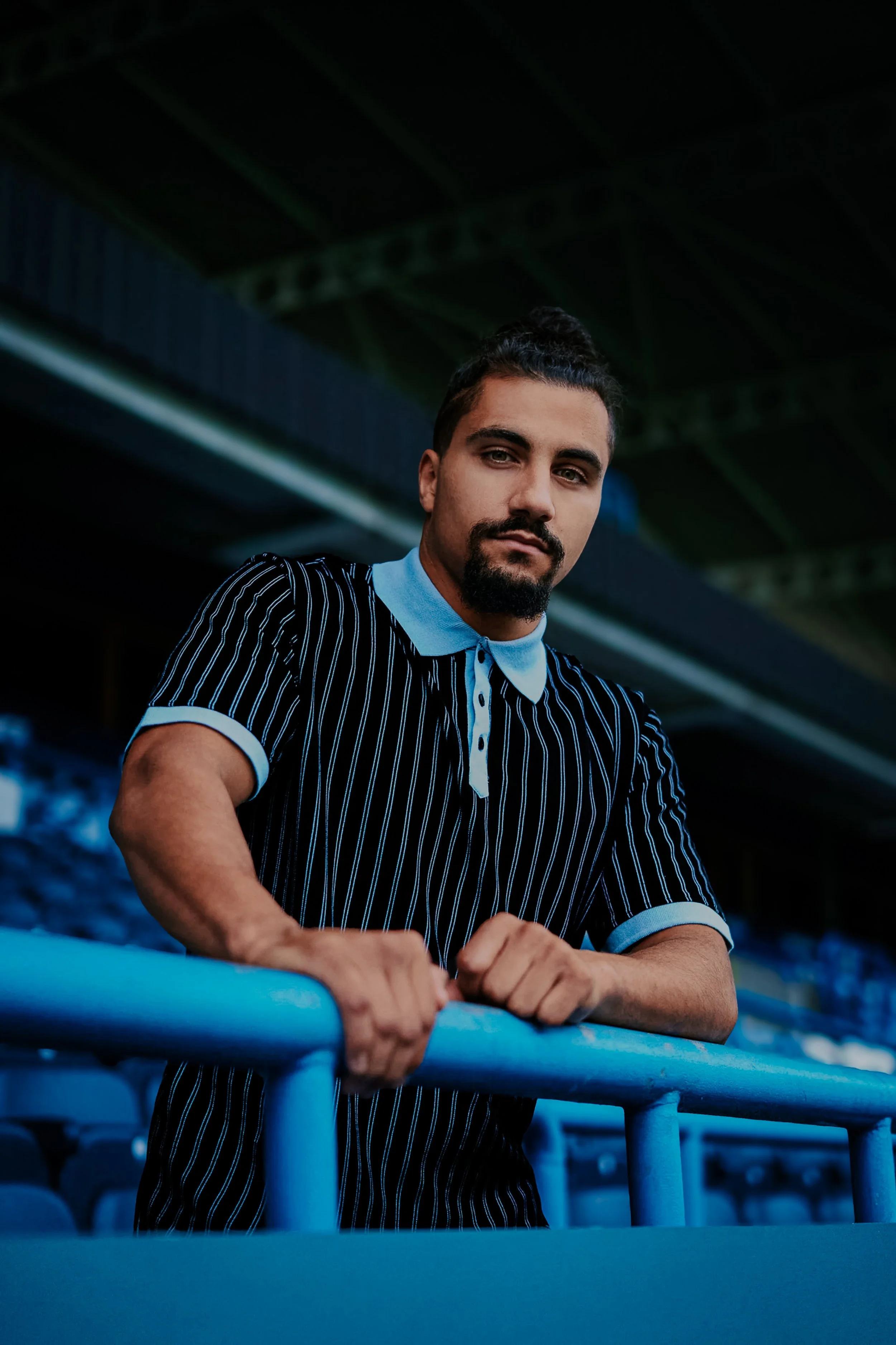 A man with dark hair tied up in a bun, a beard, and mustache, wearing a black and white striped shirt with light blue collar and cuffs, standing in a stadium, holding onto a blue railing, with a serious expression, under a dark structure overhead.