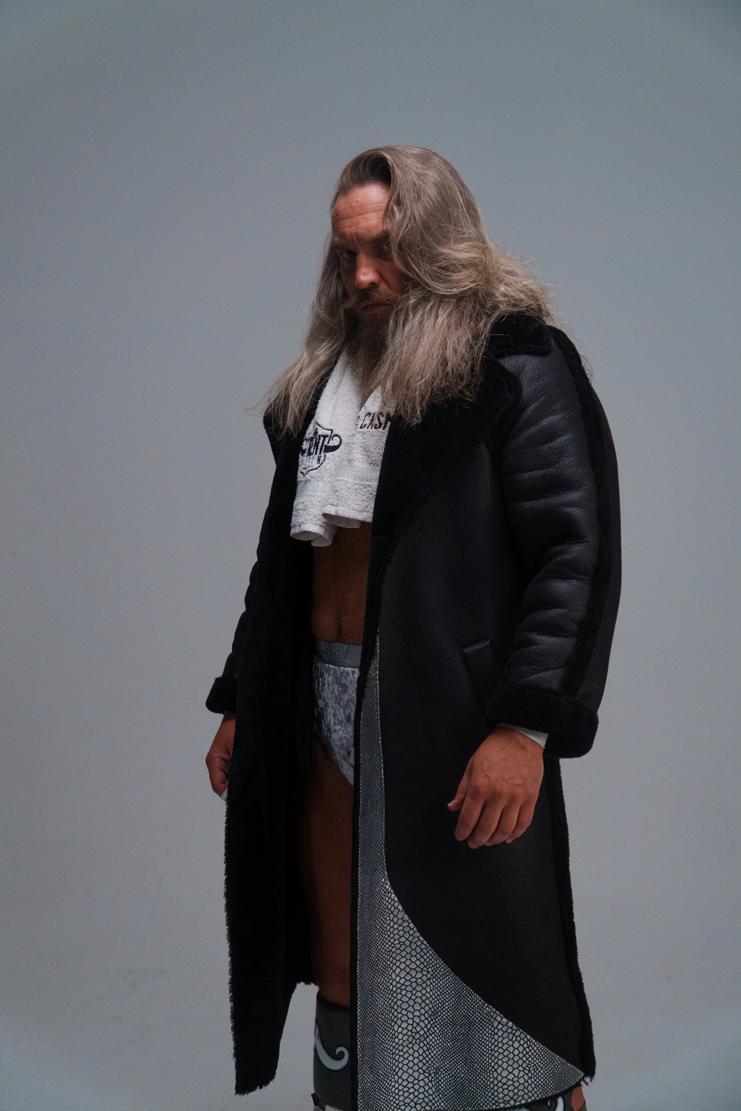 A man with long, wavy hair and a beard wearing a black coat, standing against a plain gray background.