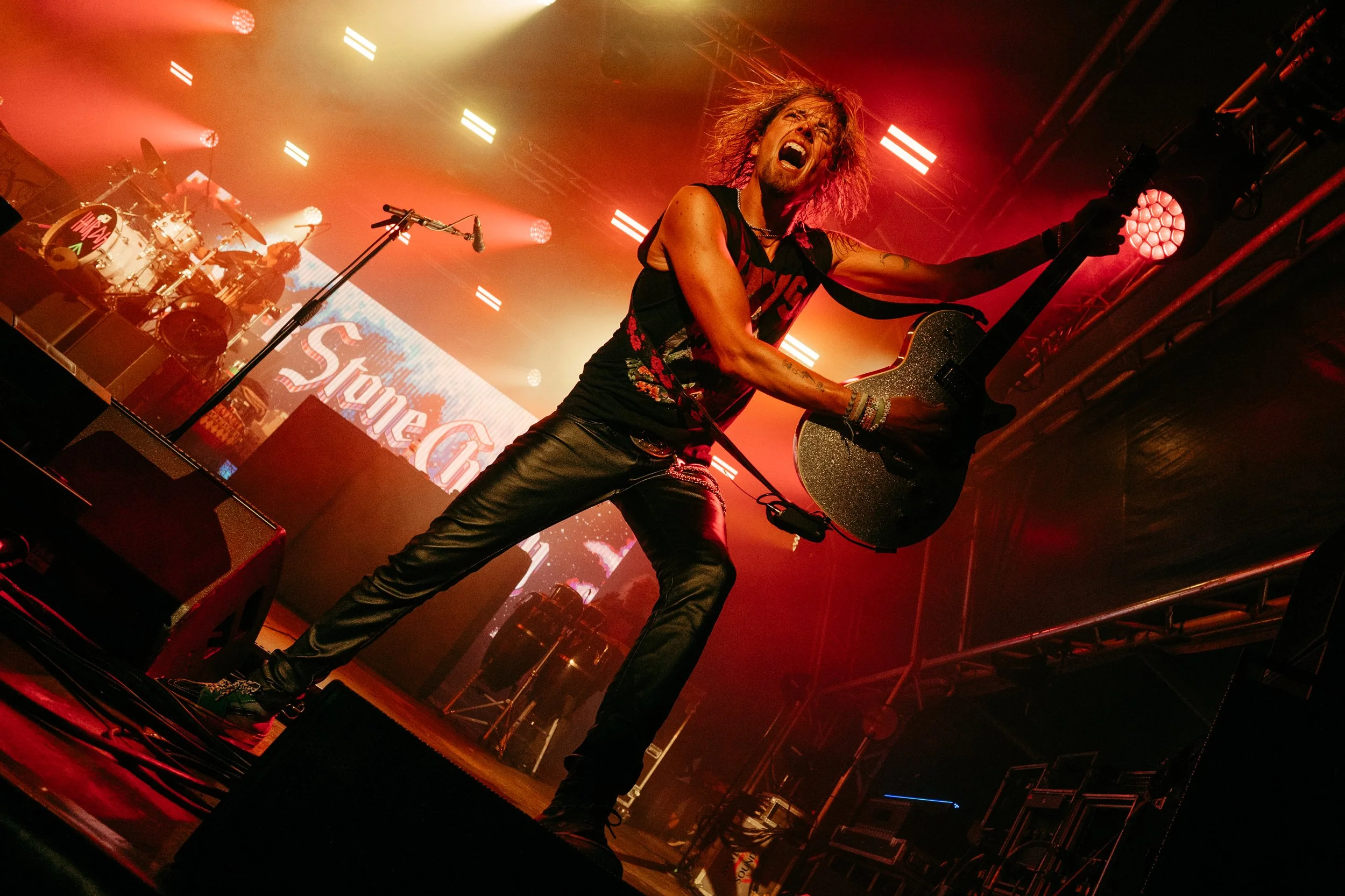 Rock musician performs on stage with an electric guitar, intense facial expression, red and yellow stage lighting, behind a drummer and a backdrop displaying part of a logo or text.