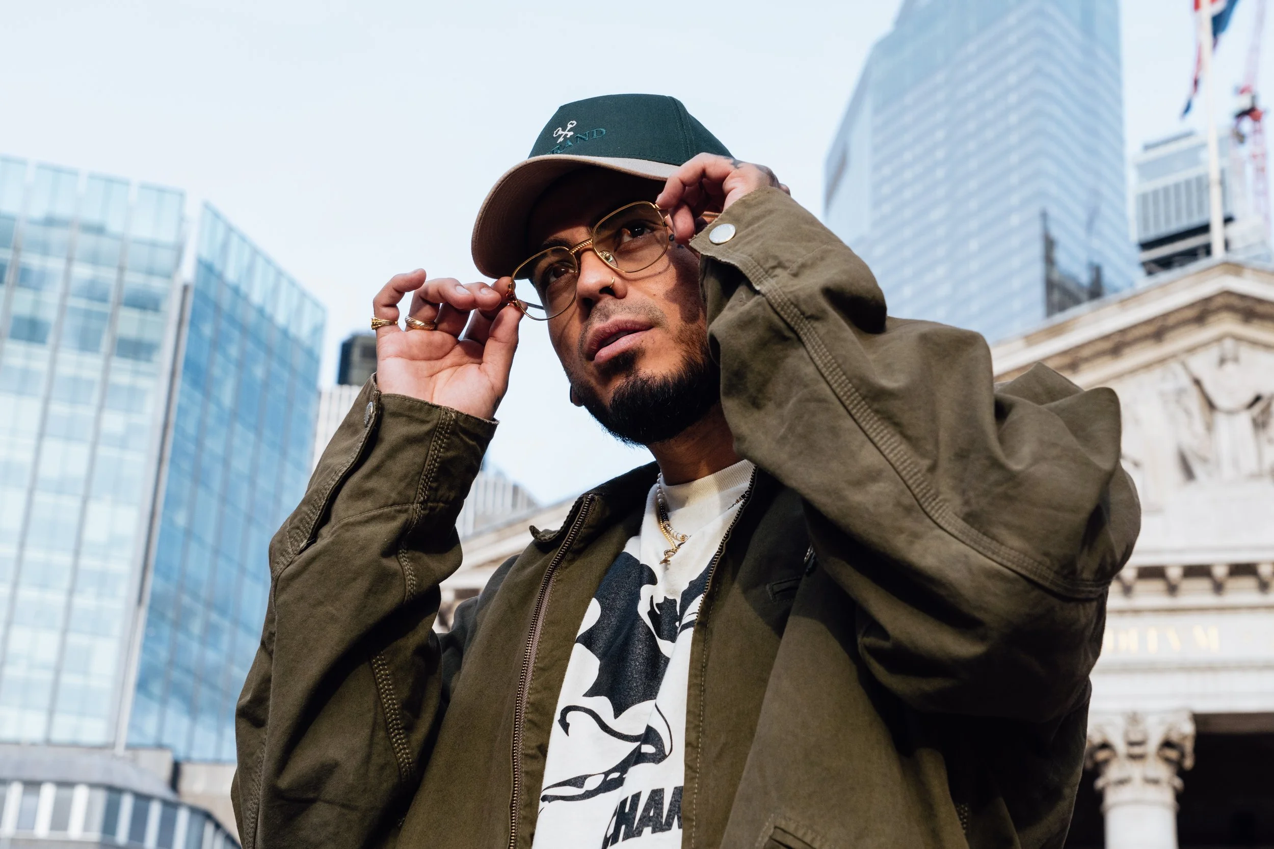 A man adjusts his glasses while standing in an urban area with tall modern buildings in the background.