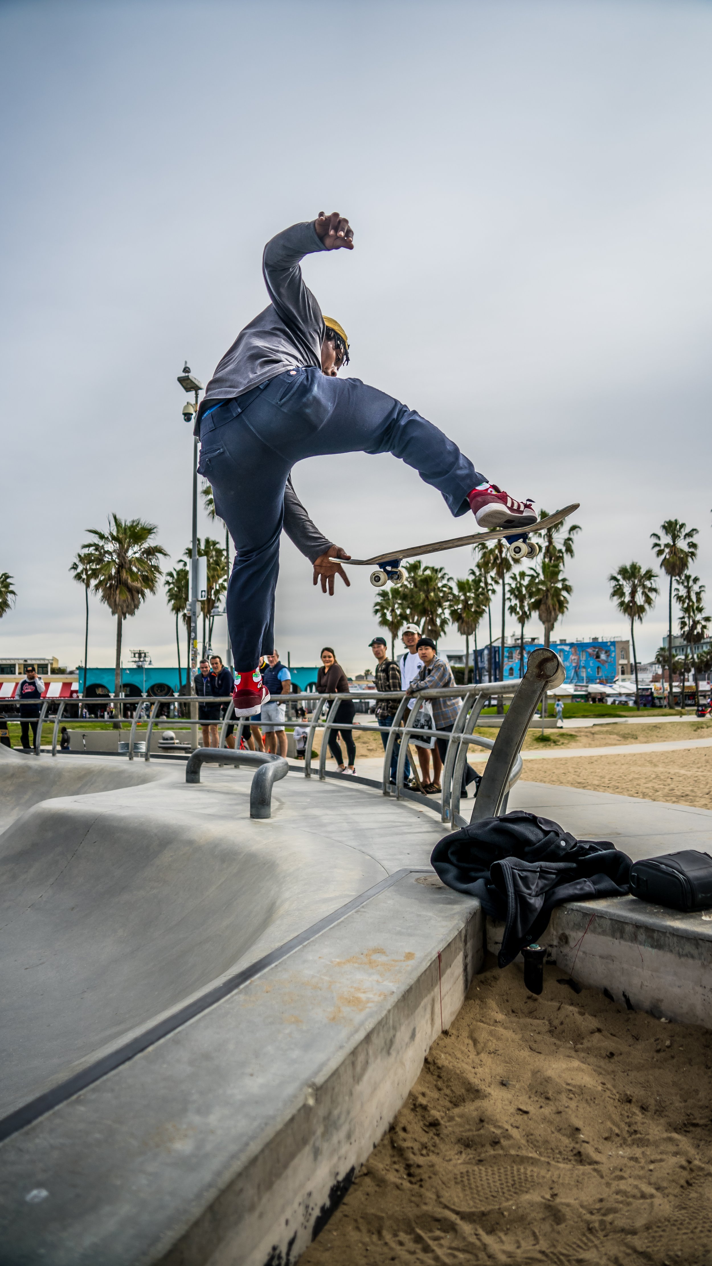 Skateboarder performing an ollie at a skate park with onlookers and palm trees in the background.