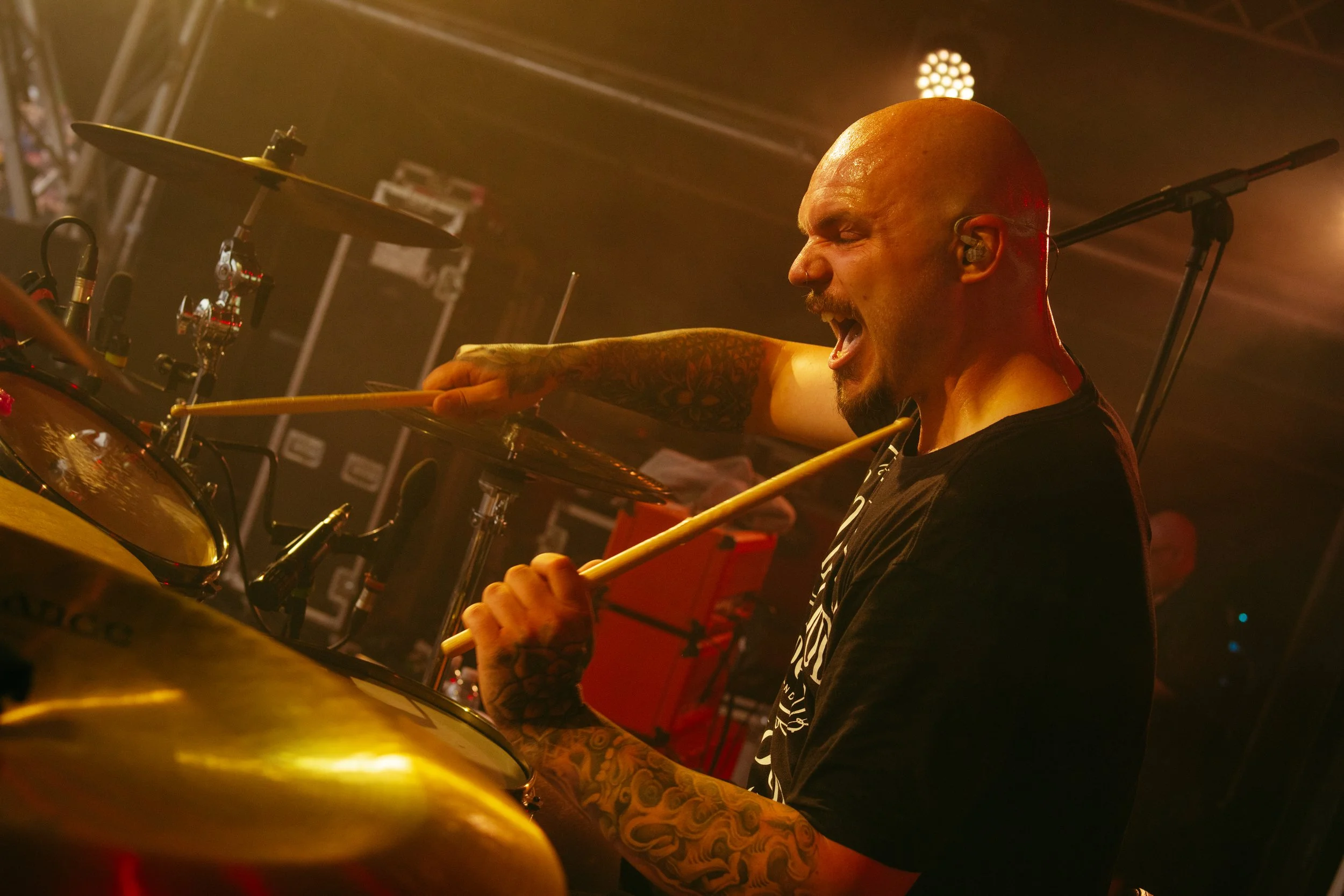 A drummer passionately plays on stage, with drumsticks in hand, surrounded by drums and cymbals, with stage lights in the background.