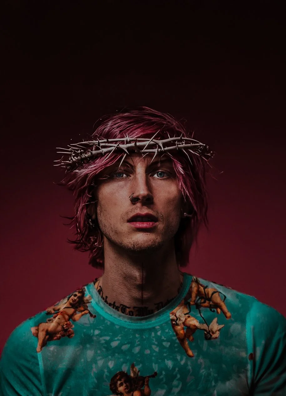 A young man with pink hair, piercing blue eyes, and a nose ring wearing a turquoise shirt with a cherub print, against a dark, gradient background.