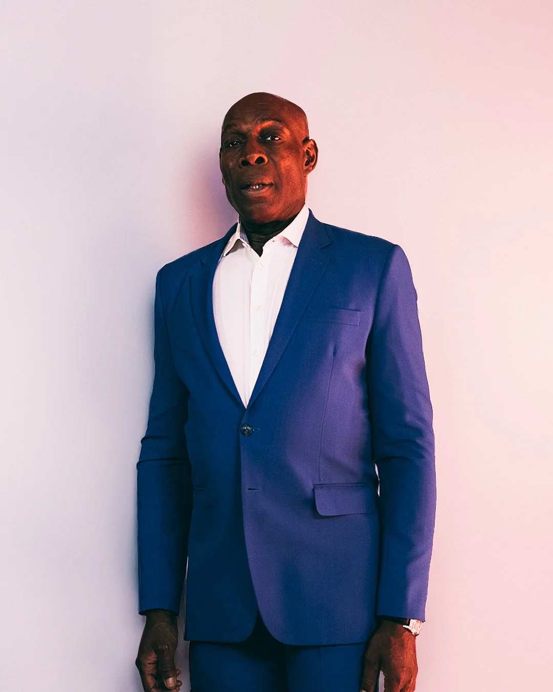 A man in a blue suit with a white shirt standing against a plain light-colored wall.