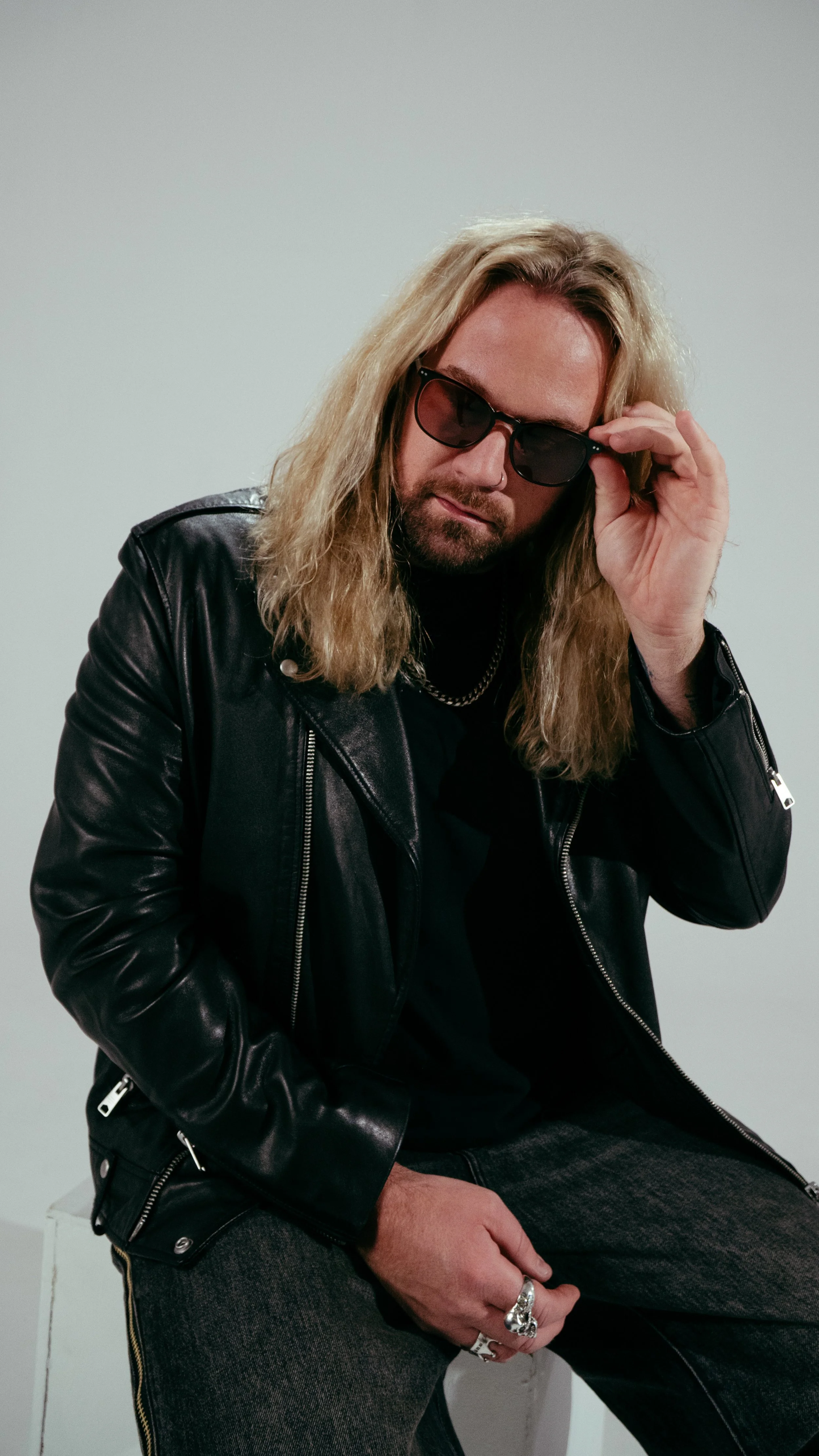 A man with long blonde hair wearing sunglasses, a black leather jacket, and dark jeans, sitting against a plain light gray wall.