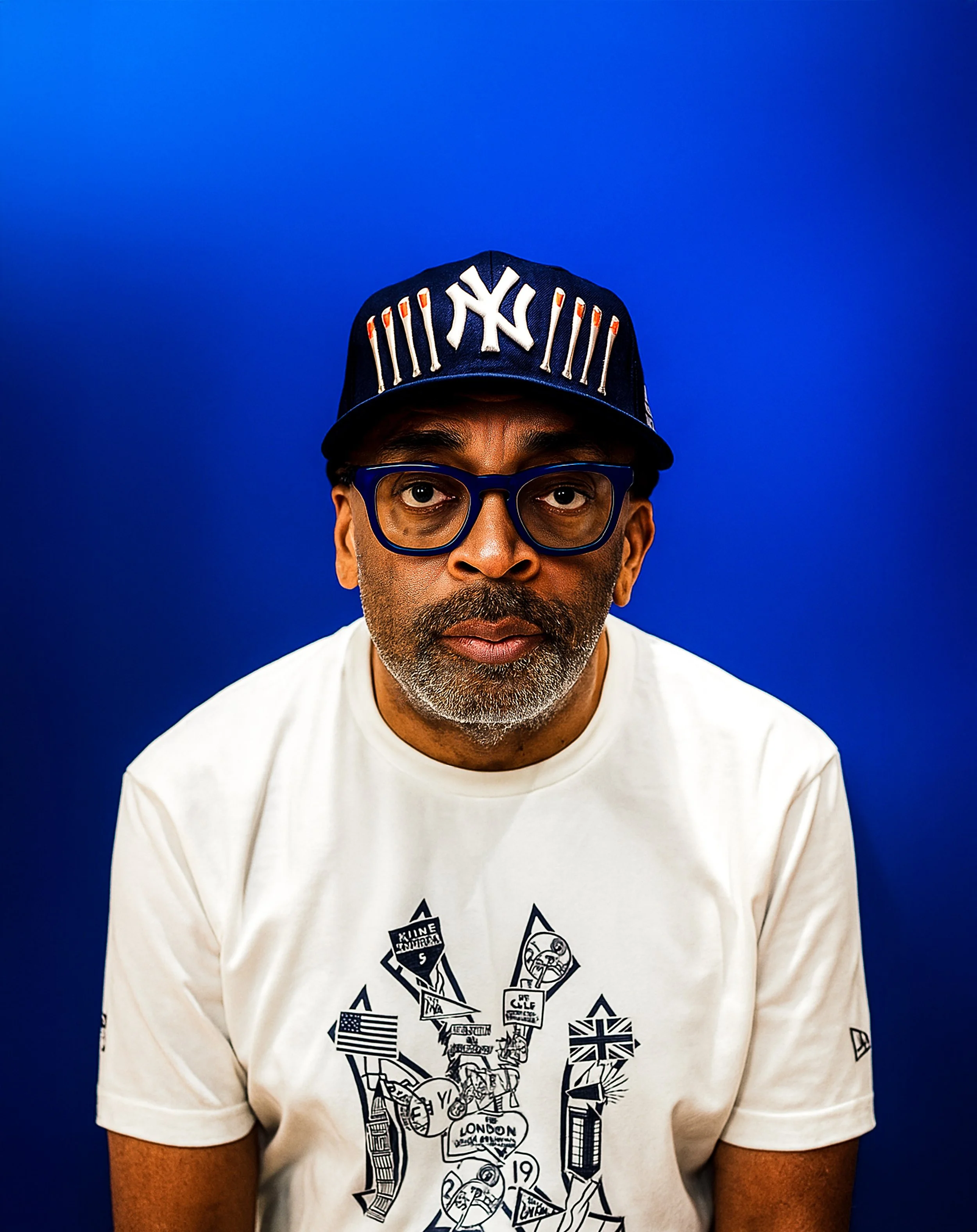 A man with glasses wearing a New York Yankees cap and a white T-shirt with graphic illustrations, standing against a solid blue background.