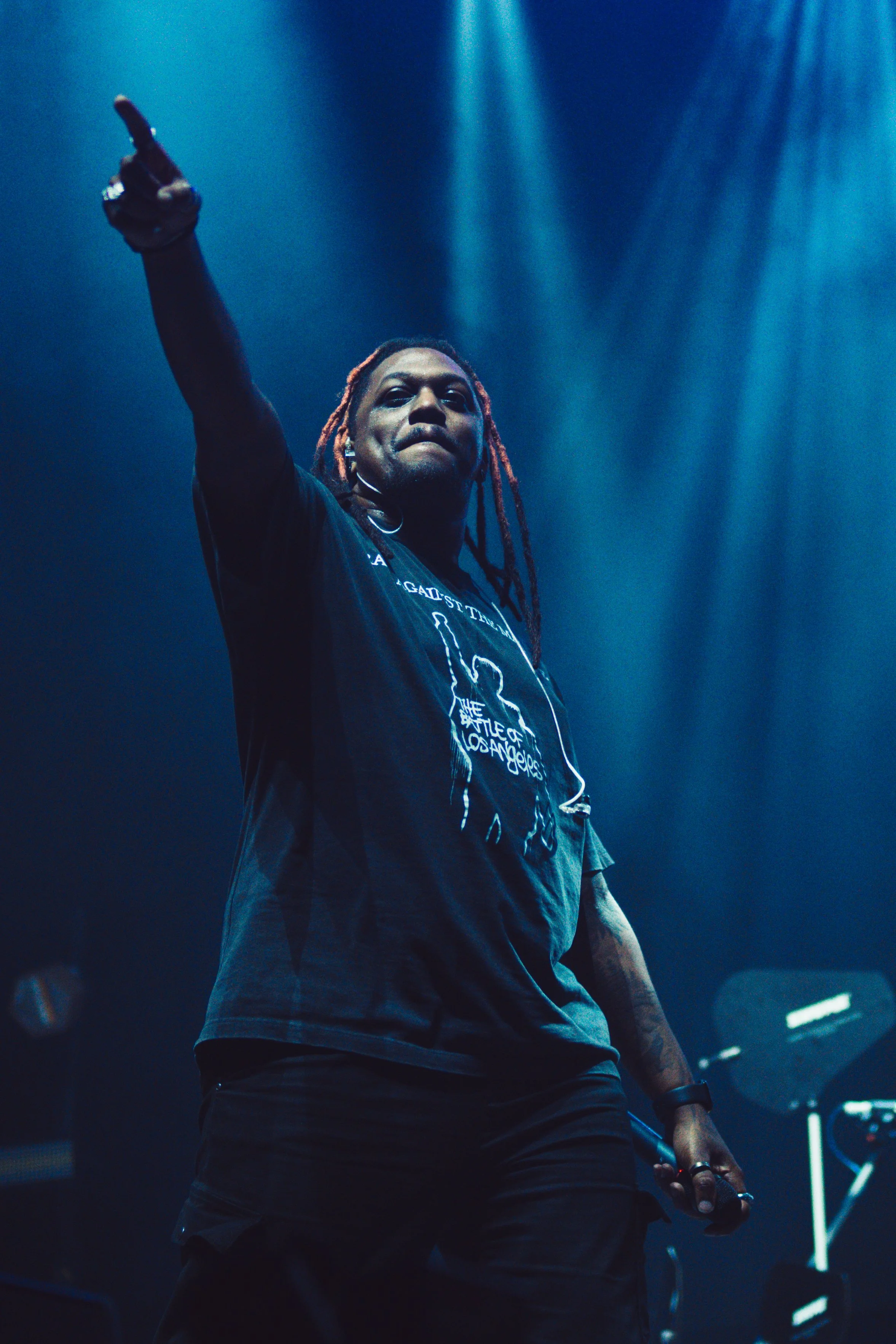 A performer with dreadlocks on stage, wearing a black t-shirt, pointing upward with stage lights shining down.