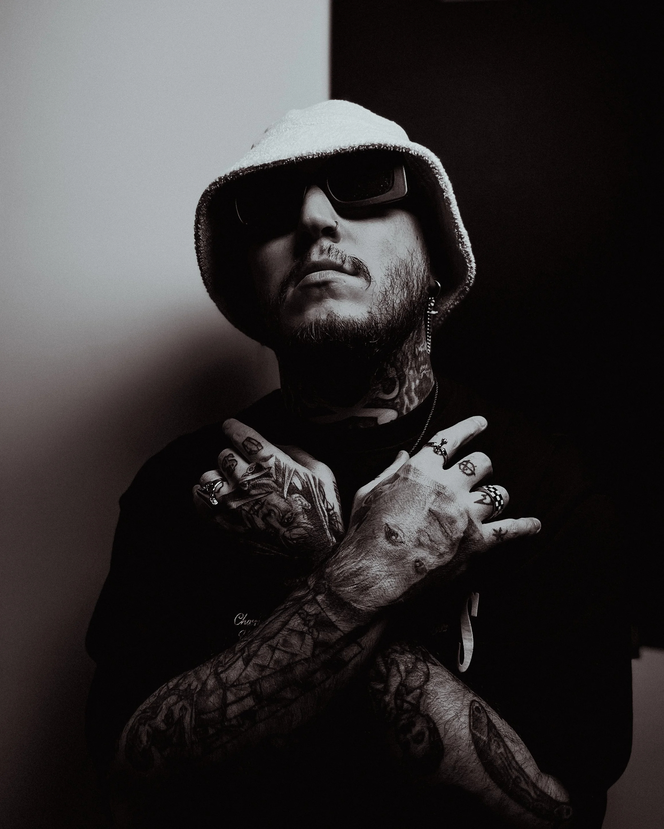 A man with tattoos, wearing sunglasses, a hoodie, and earrings, poses against a black and white background, with one arm crossed over his chest and the other arm resting on it.