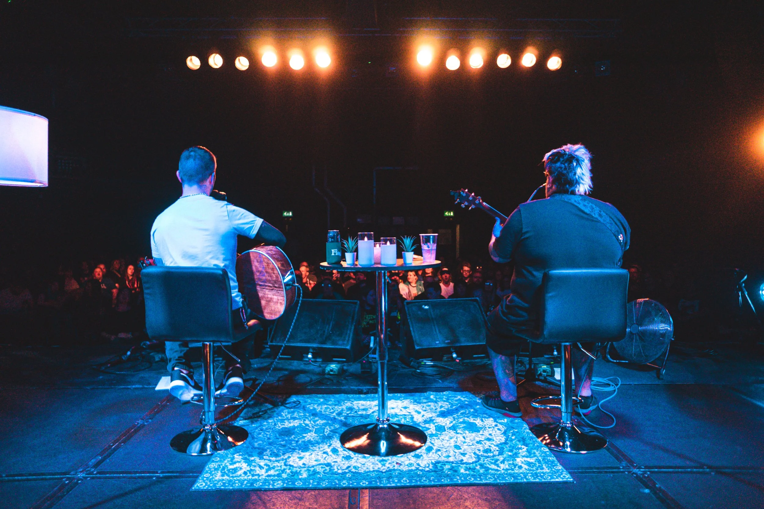 Two musicians performing on stage with audience watching, illuminated by stage lights, with candles and small potted plants on a table in front.