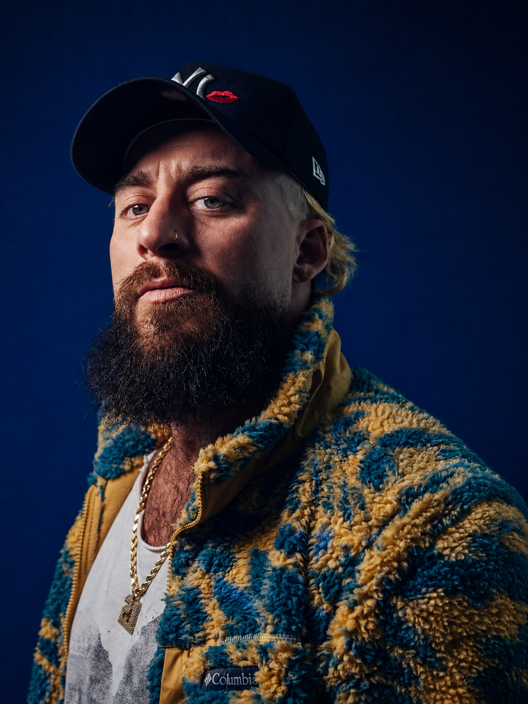 Portrait of a man with a beard wearing a black baseball cap, a yellow and blue textured fleece jacket, a white T-shirt, gold chains, and a nose ring, set against a dark blue background.