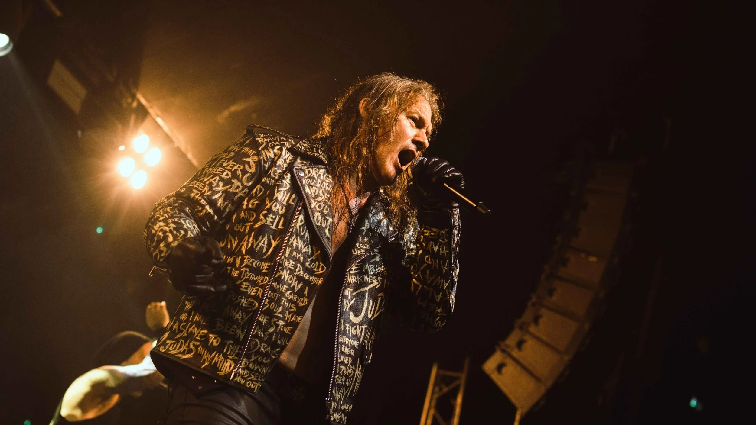 A male singer with long curly hair wearing a black jacket covered in yellow writing, singing into a microphone on stage with bright stage lights behind him.