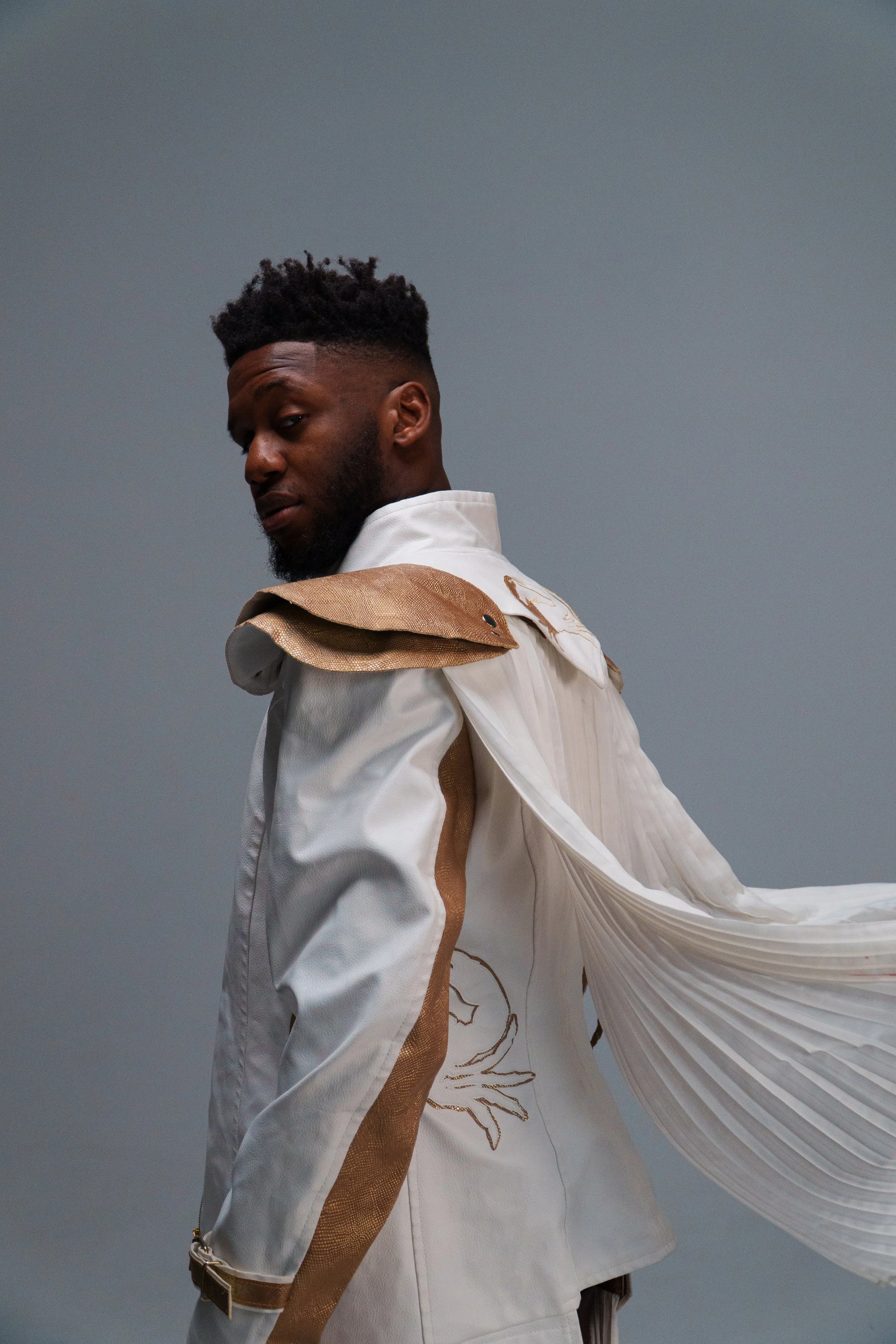 A man in a white and gold jacket with pleated cape details poses against a plain gray background.