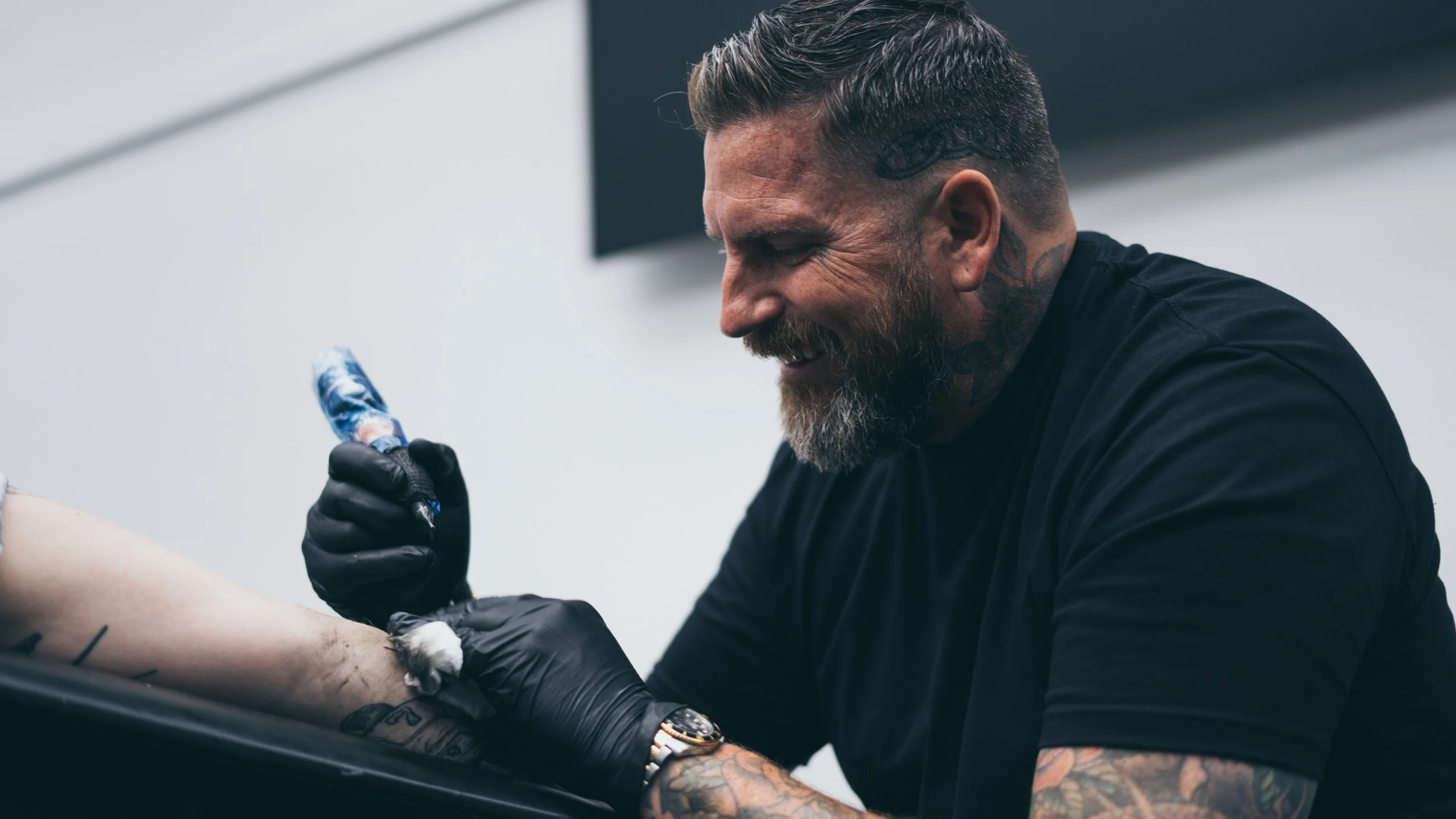 A tattooed man with a beard and slicked-back hair is tattooing a person's arm in a studio with white walls.
