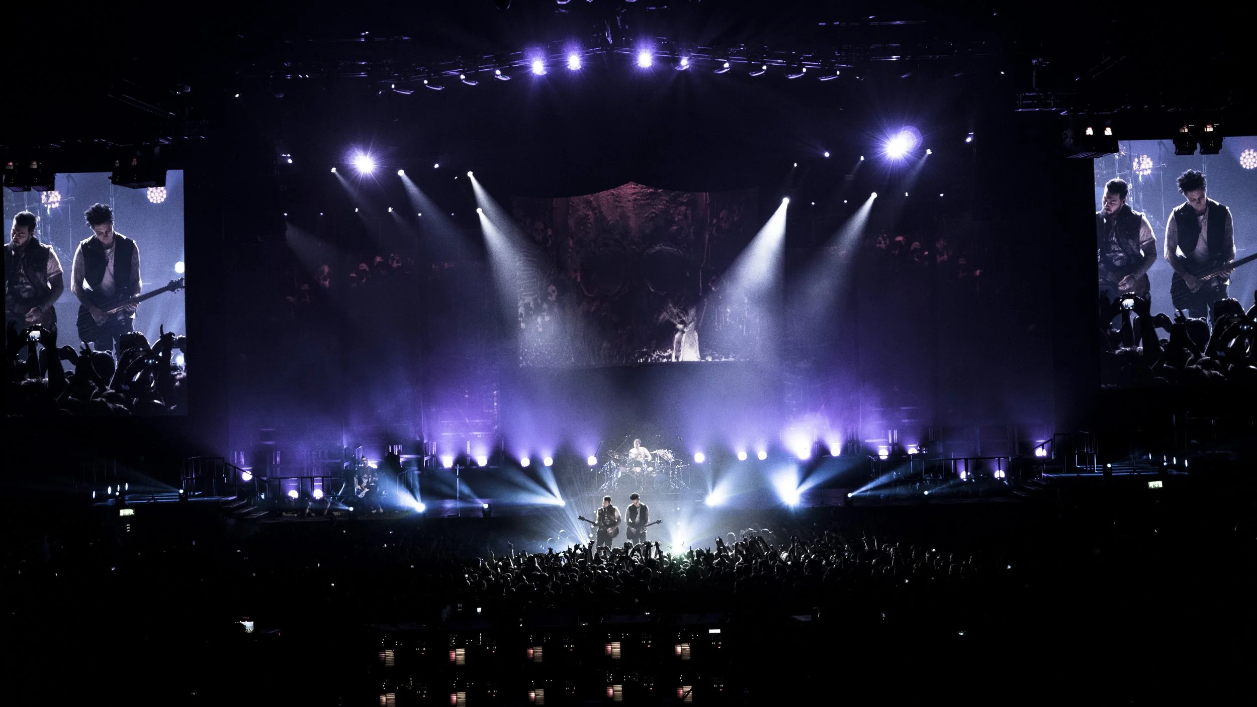 Concert stage illuminated with purple lights, musicians performing, large screens on both sides showing close-ups of guitarists, audience with raised hands in the foreground.