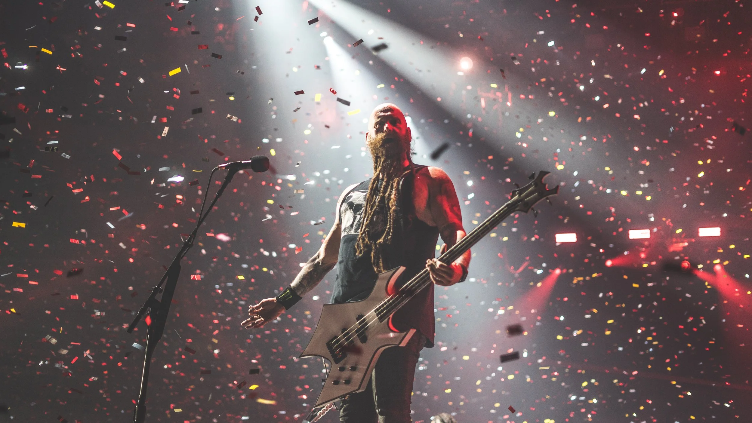 A musician with long dreadlocks and a beard plays an electric bass guitar on stage during a concert, illuminated by a spotlight with colorful confetti falling around him.