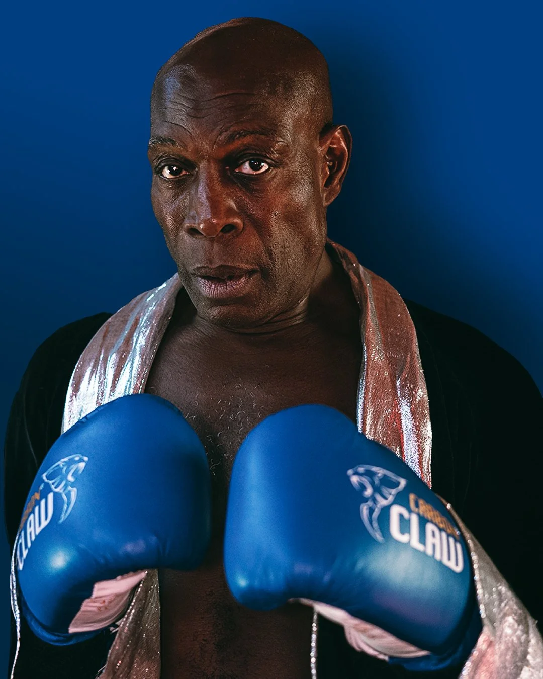 A boxer wearing blue focus mitts, with a serious expression, against a blue background.