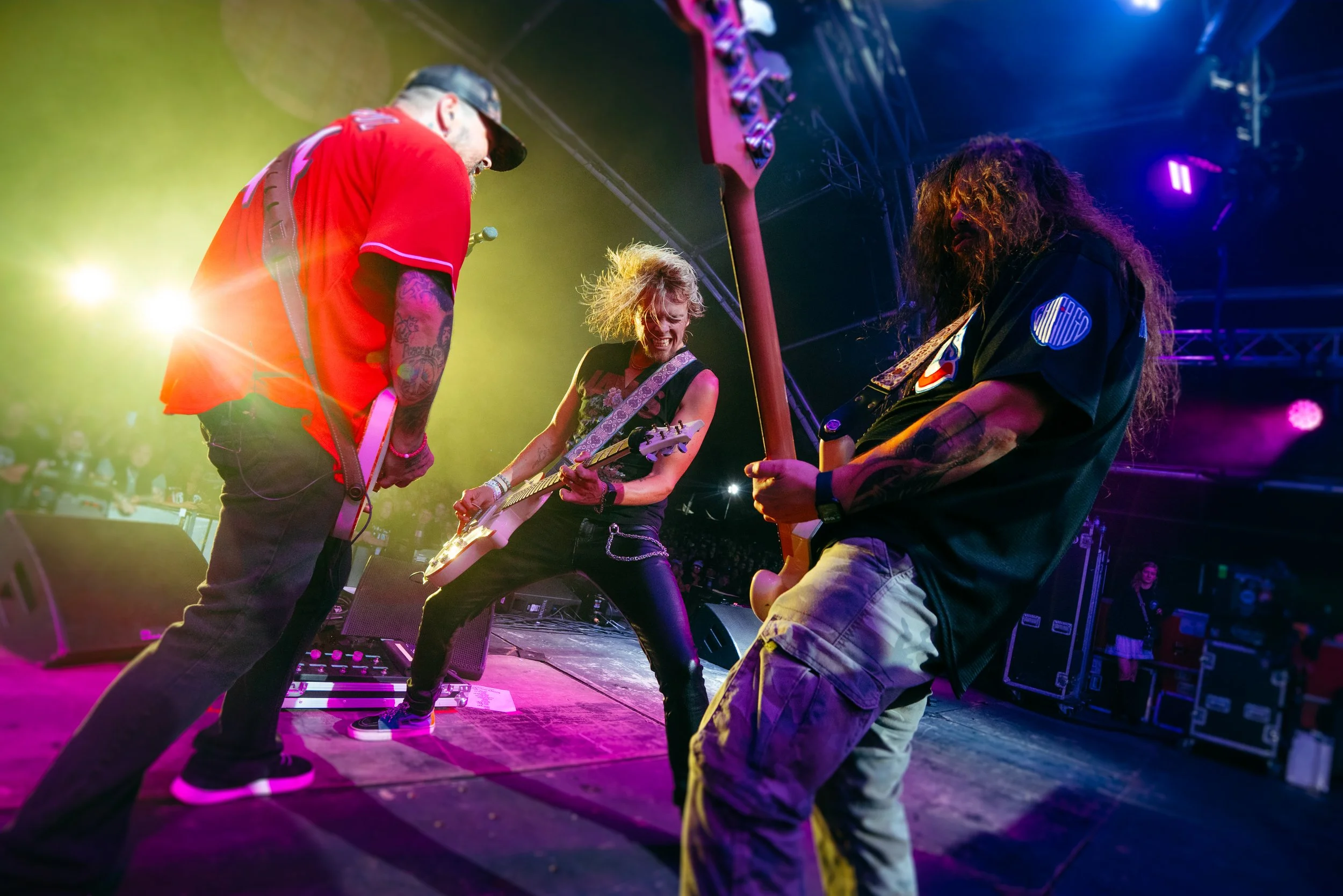 Three musicians perform on stage with colorful lighting; two men and one woman are playing electric guitars with intense expressions.