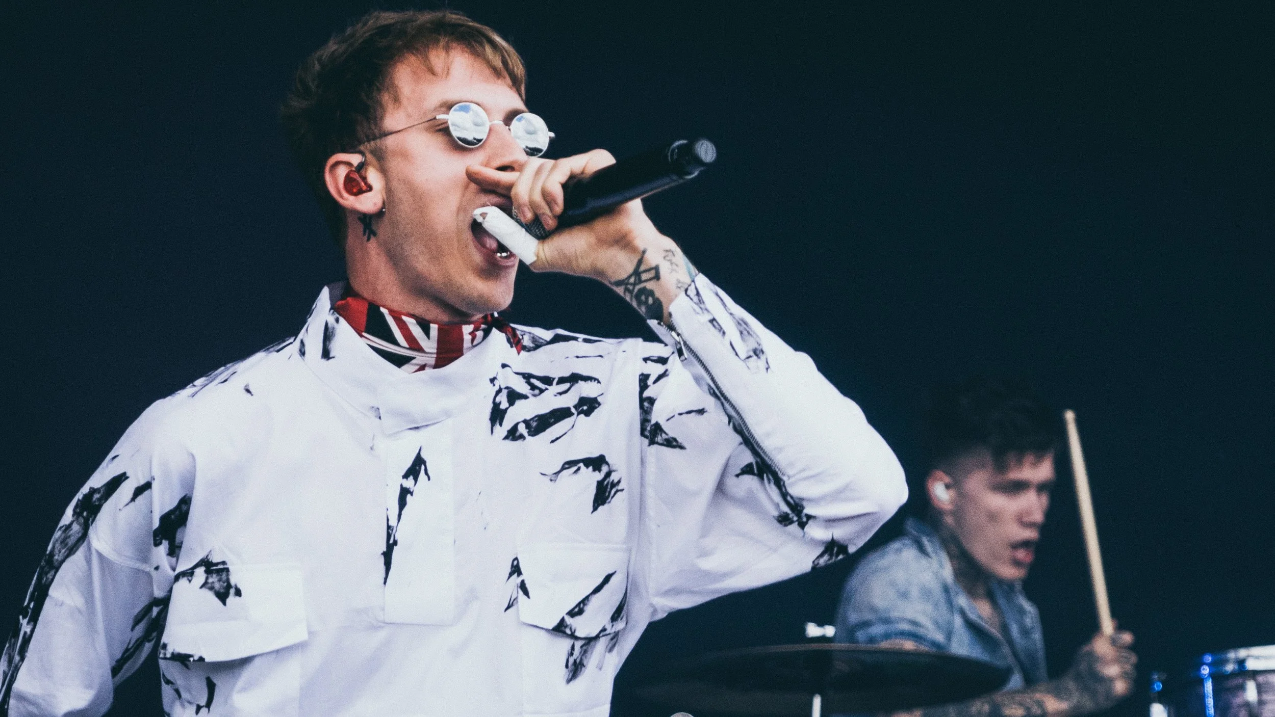 A male singer performs on stage, holding a microphone and wearing glasses with small round lenses, a white patterned jacket, and a patterned neck scarf. There is a drummer in the background playing drums.