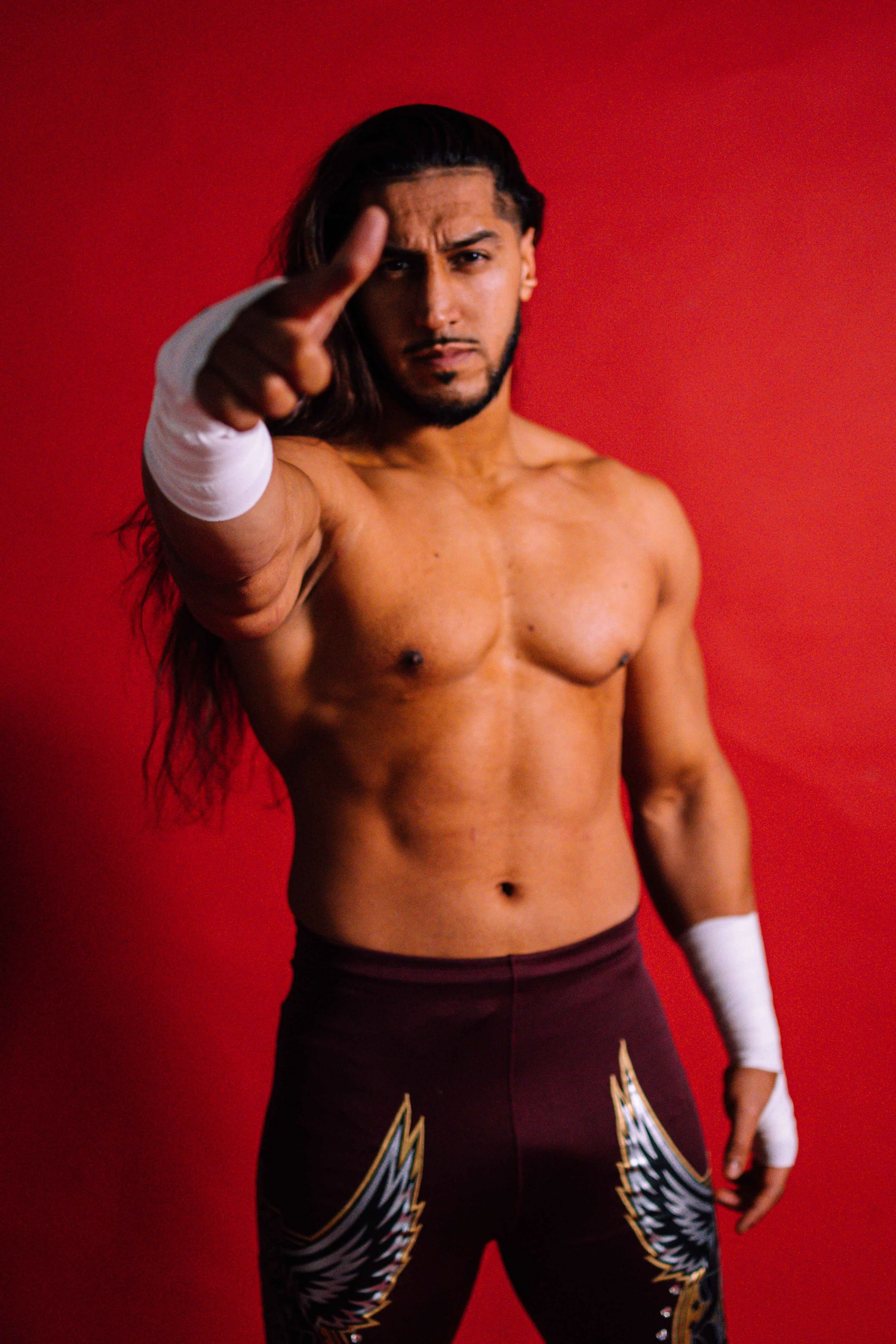 A shirtless man with long dark hair and a serious expression, pointing at the camera with one finger, against a red background. He has a white bandage on his left wrist and black athletic pants with eagle wing designs.